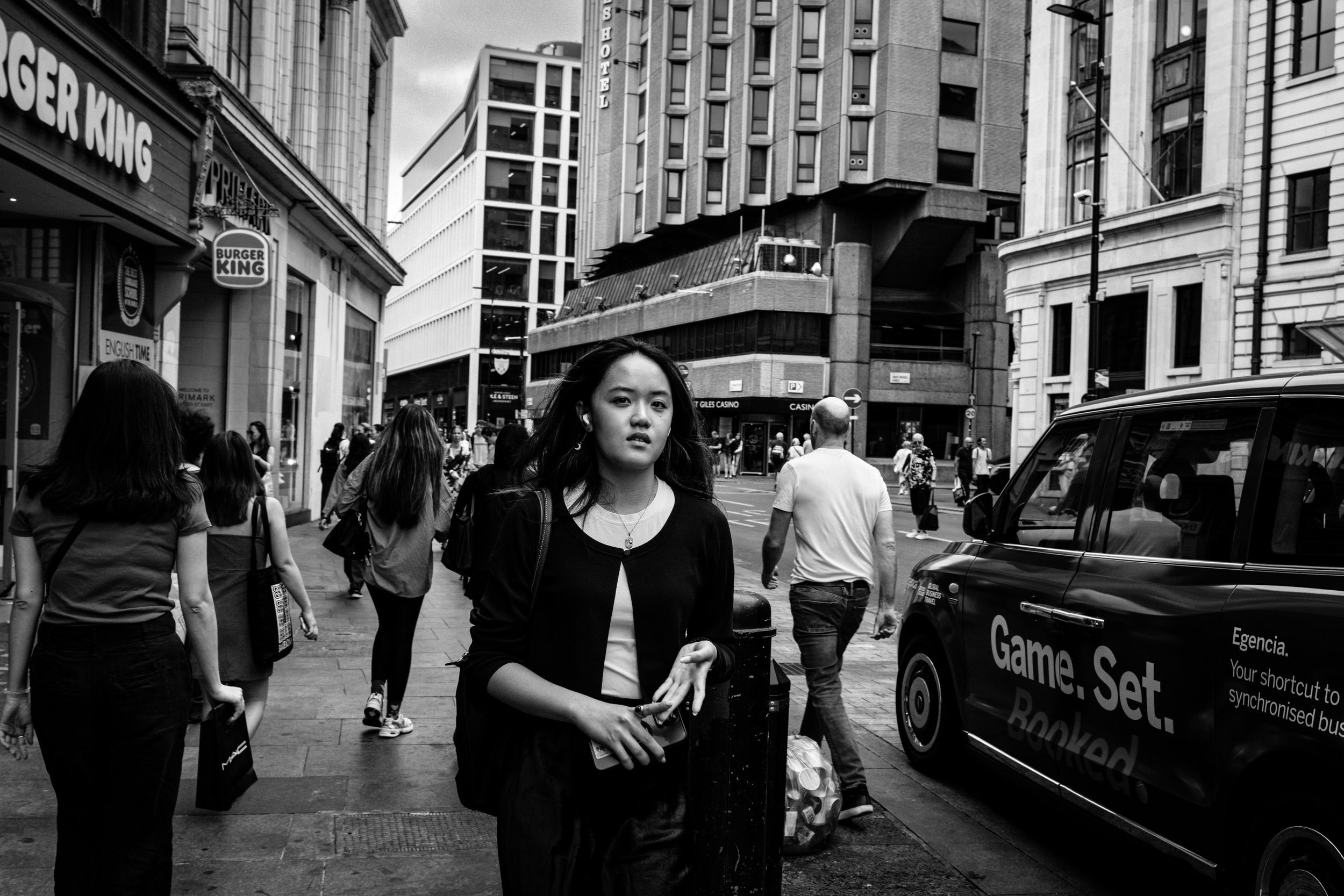A black-and-white photo of a busy city street with pedestrians, tall buildings, and a Burger King sign visible on the left side. In the foreground, a young woman with long dark hair and earrings stands, holding a shopping bag.