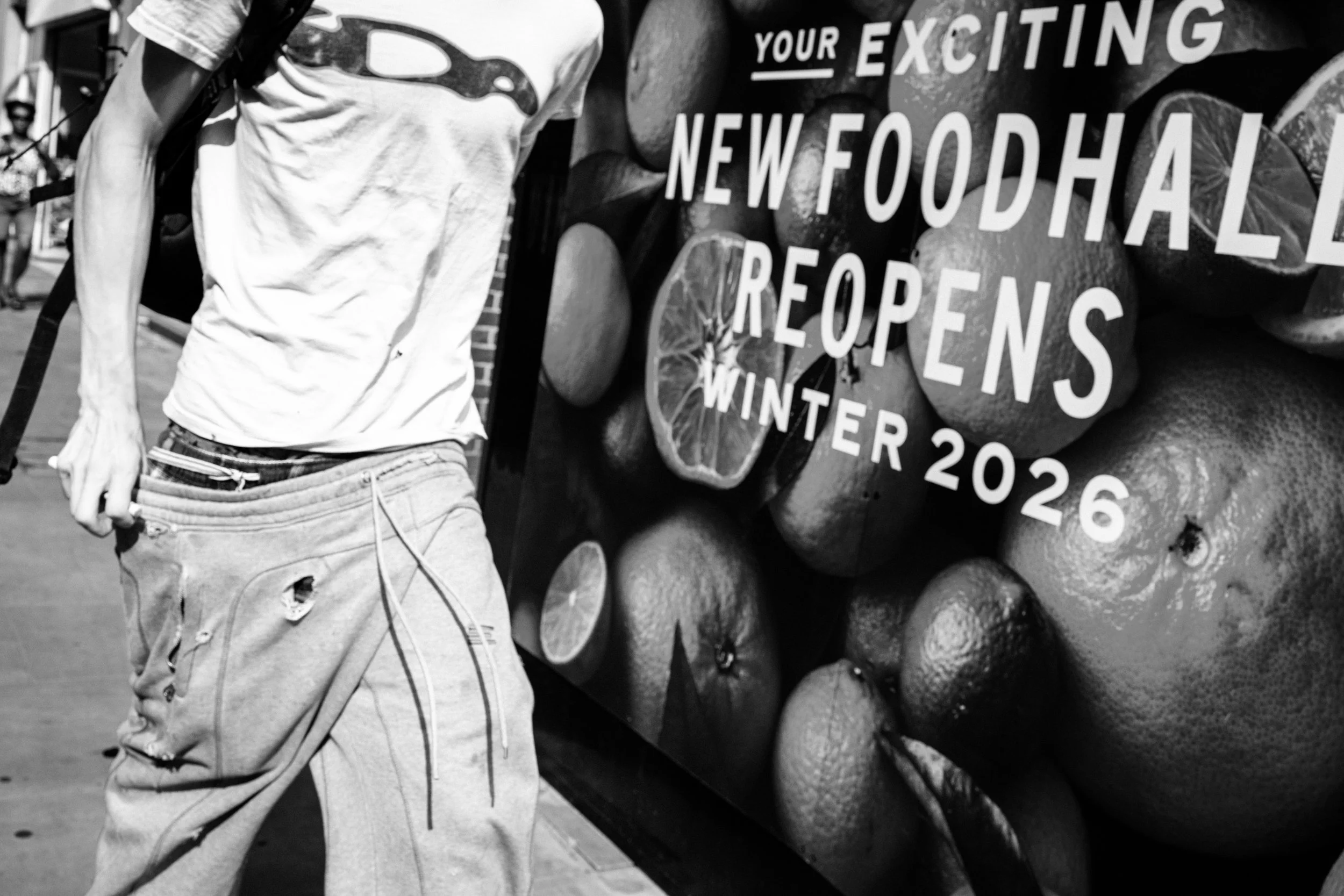 A person wearing a white T-shirt with a logo and ripped jeans standing next to a display of fresh citrus fruits with a sign that reads 'Your exciting new food hall reopening winter 2026.'