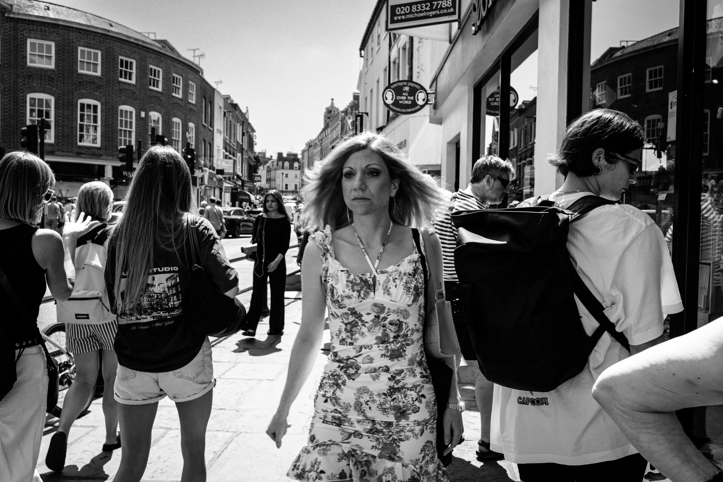 A black-and-white photo of a busy street scene with multiple pedestrians walking on a sidewalk, some are talking to each other, and others are looking at shops. One woman in a floral dress is walking towards the camera, while others are on either side of her, with some partially cut off in the frame. Shops and buildings line the street, and there are signs hanging from the buildings.
