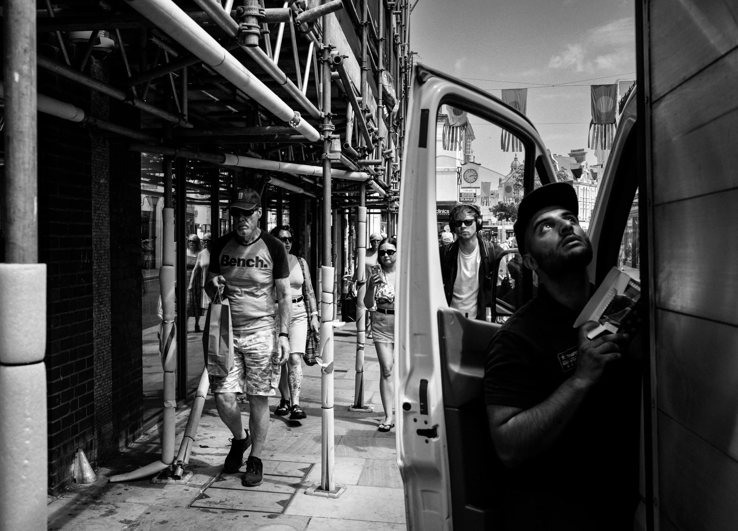 Black and white photo of pedestrians walking on a city sidewalk near a scaffolding structure, with a person operating a small vehicle on the right side of the image, seen from inside the vehicle. Buildings and a clock tower are visible in the background.