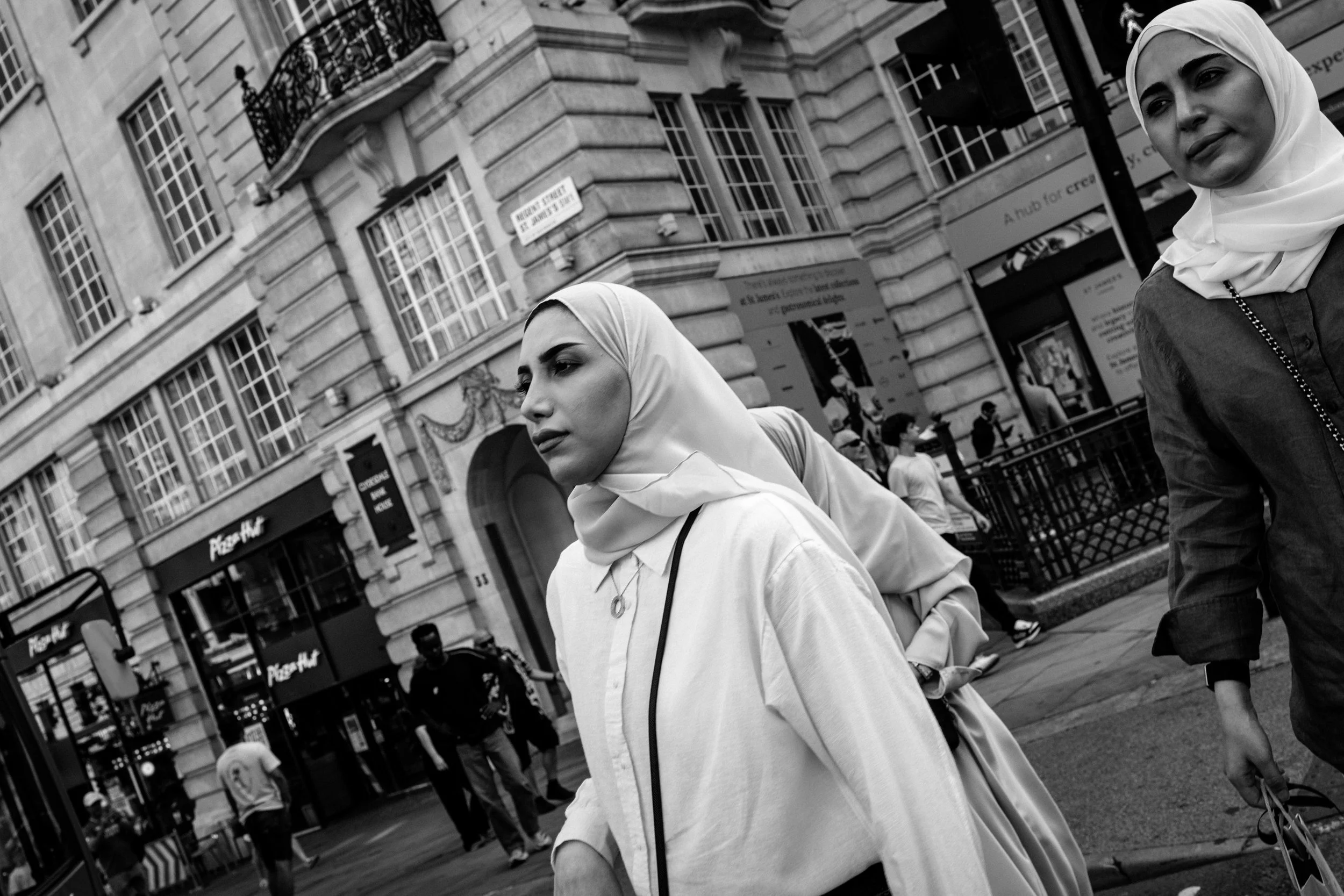 Two women wearing headscarves walking on a busy city street surrounded by buildings, pedestrians, and storefronts.