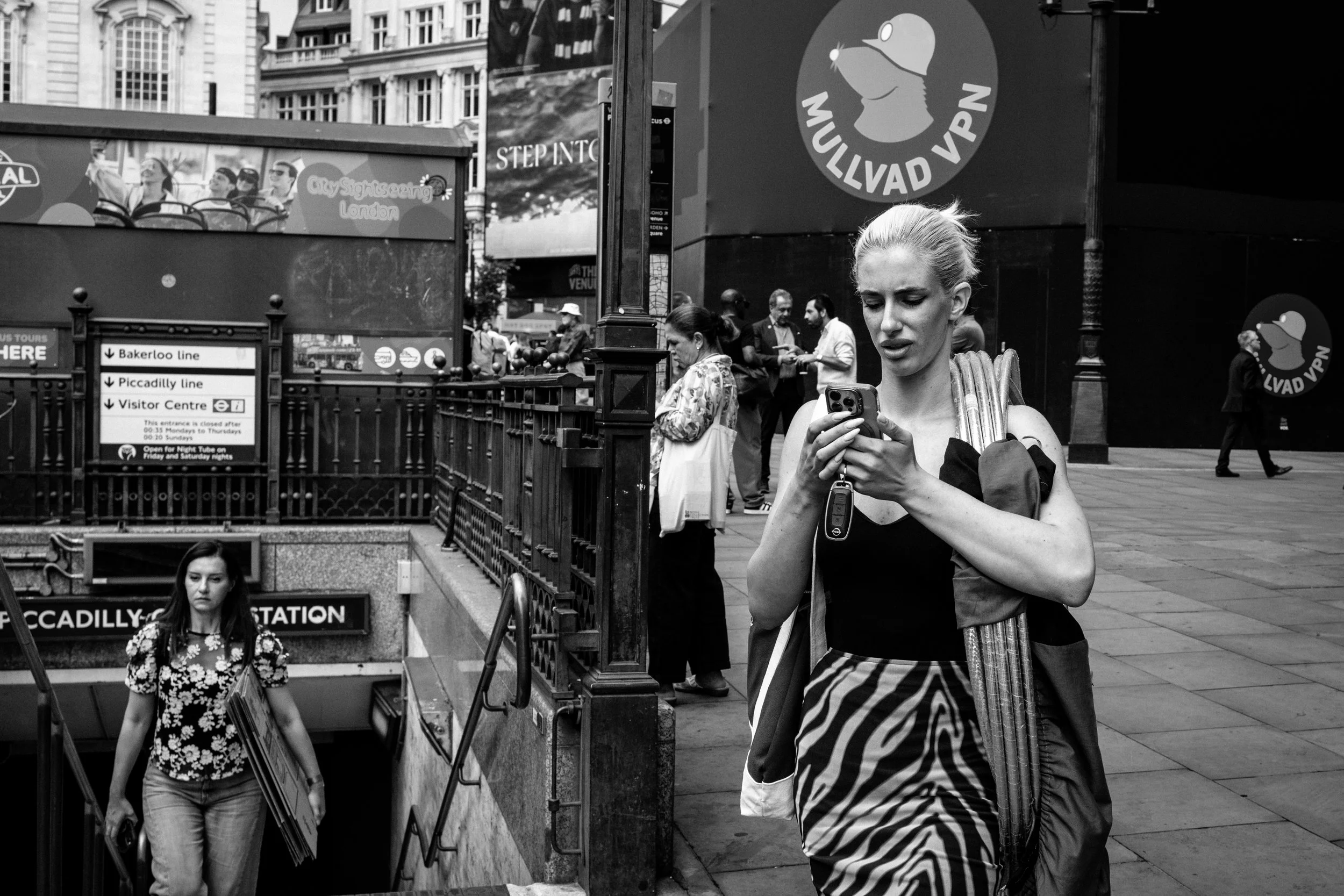 A black and white photo of a busy city street with people exiting an underground station. A woman with blonde hair in a bun is looking at her phone and holding an umbrella. Other pedestrians are walking by; one woman is holding a picture frame. There are signs for the Piccadilly line, visitor center, and a large sign with a duck logo for MULLVAD VPN.