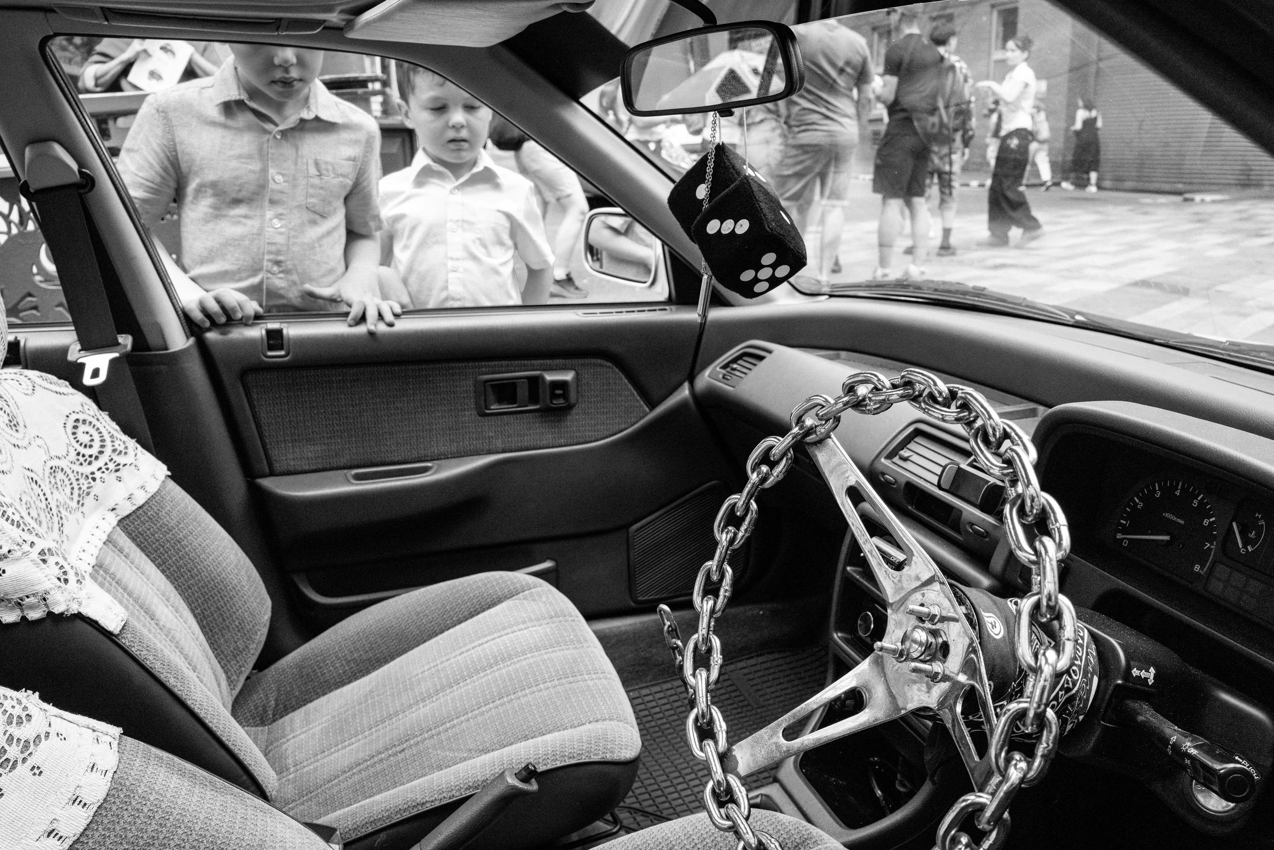 Inside view of a car trunk with chained bike, seen from the front seats. Two boys are looking inside from outside the vehicle, with people and a building in the background.