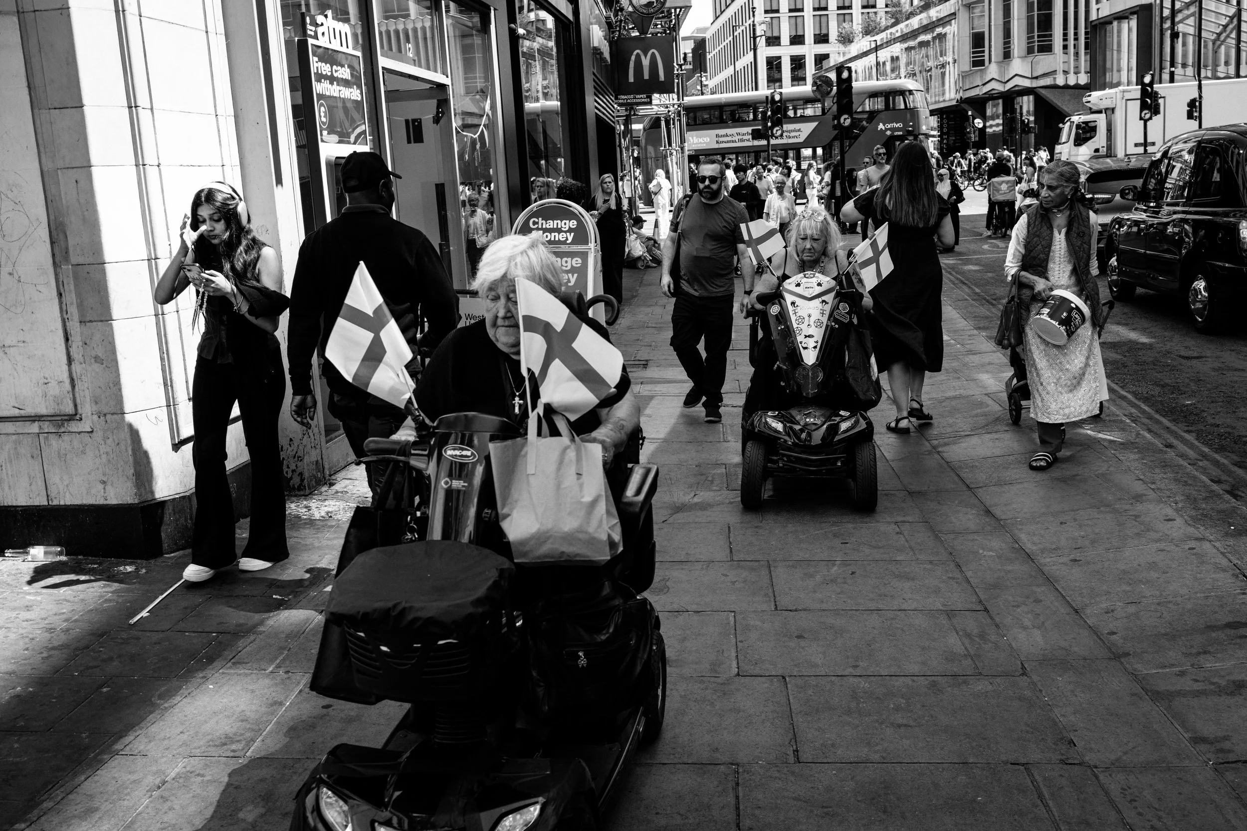 Black and white photo of a busy city sidewalk with people walking, some carrying flags, and a small vehicle with flags on a city street. There are storefronts, traffic lights, and tall buildings in the background.