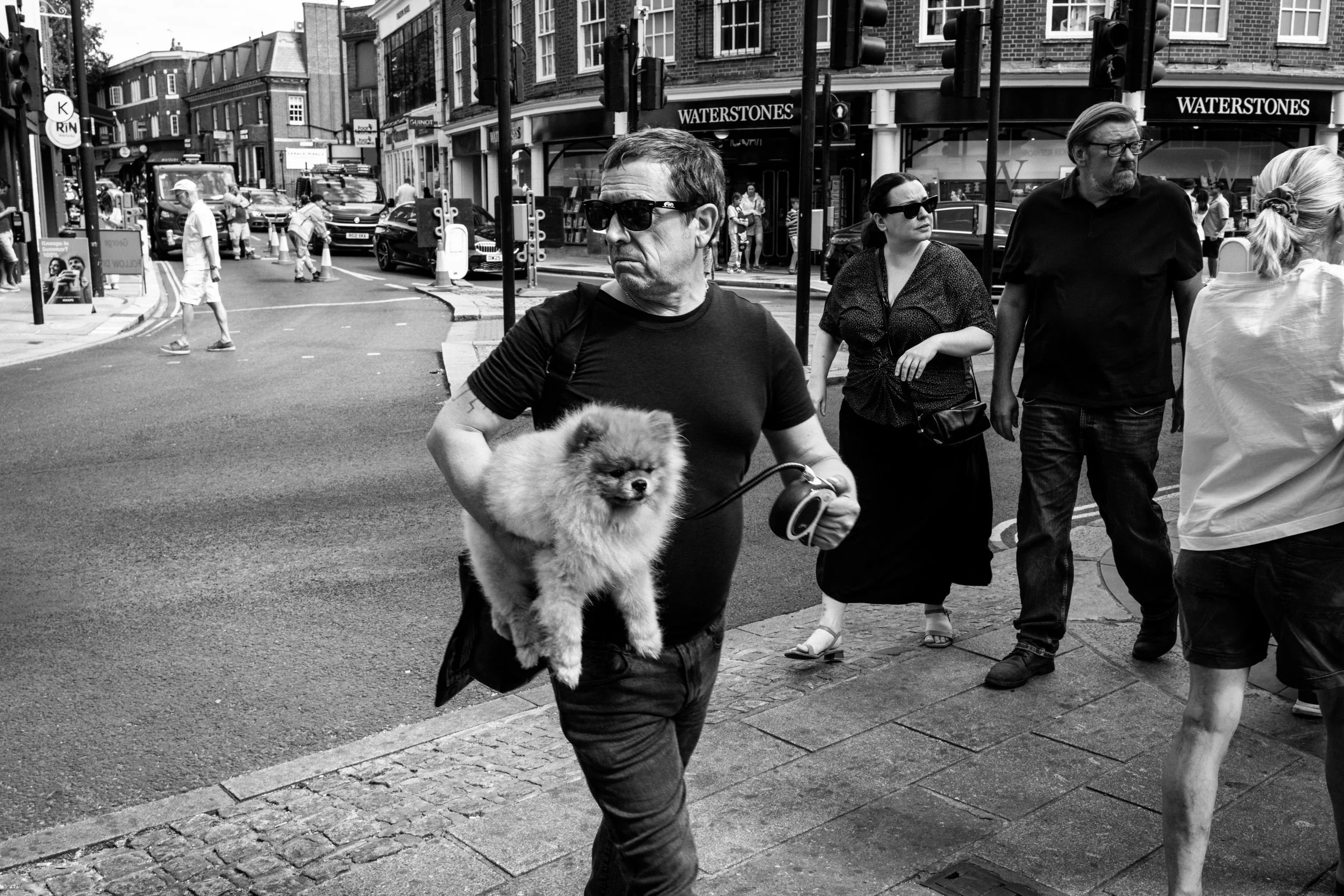 A black and white photo of people crossing a city street, including a man holding a small fluffy dog with a leash, wearing sunglasses and a black shirt, and other pedestrians walking nearby, with storefronts and vehicles in the background.