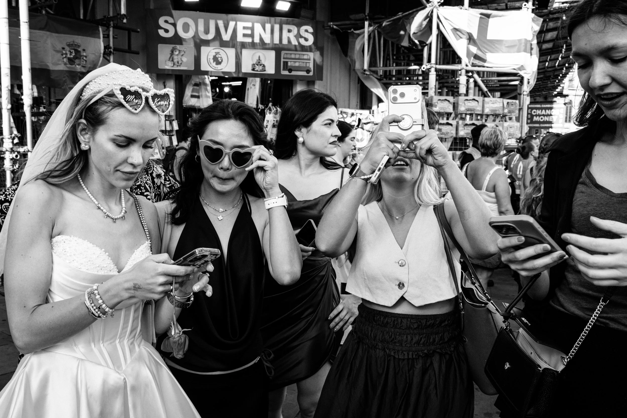 Group of women at a crowded souvenir market, some looking at their phones, others taking photos, with signs reading 'SOUVENIRS' in the background.