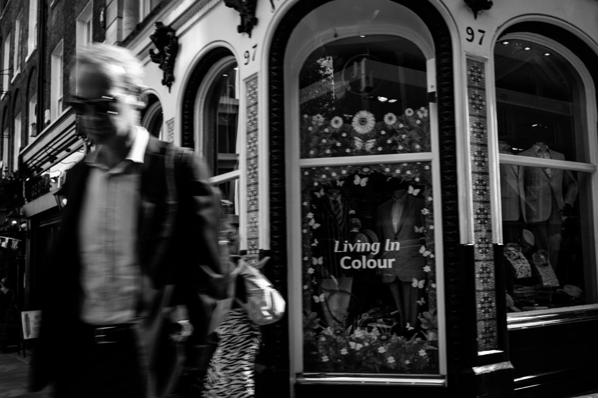 A black-and-white photo of a city street scene showing a blurred man walking past a storefront with large windows displaying floral decorations and the words "Living in Colour."