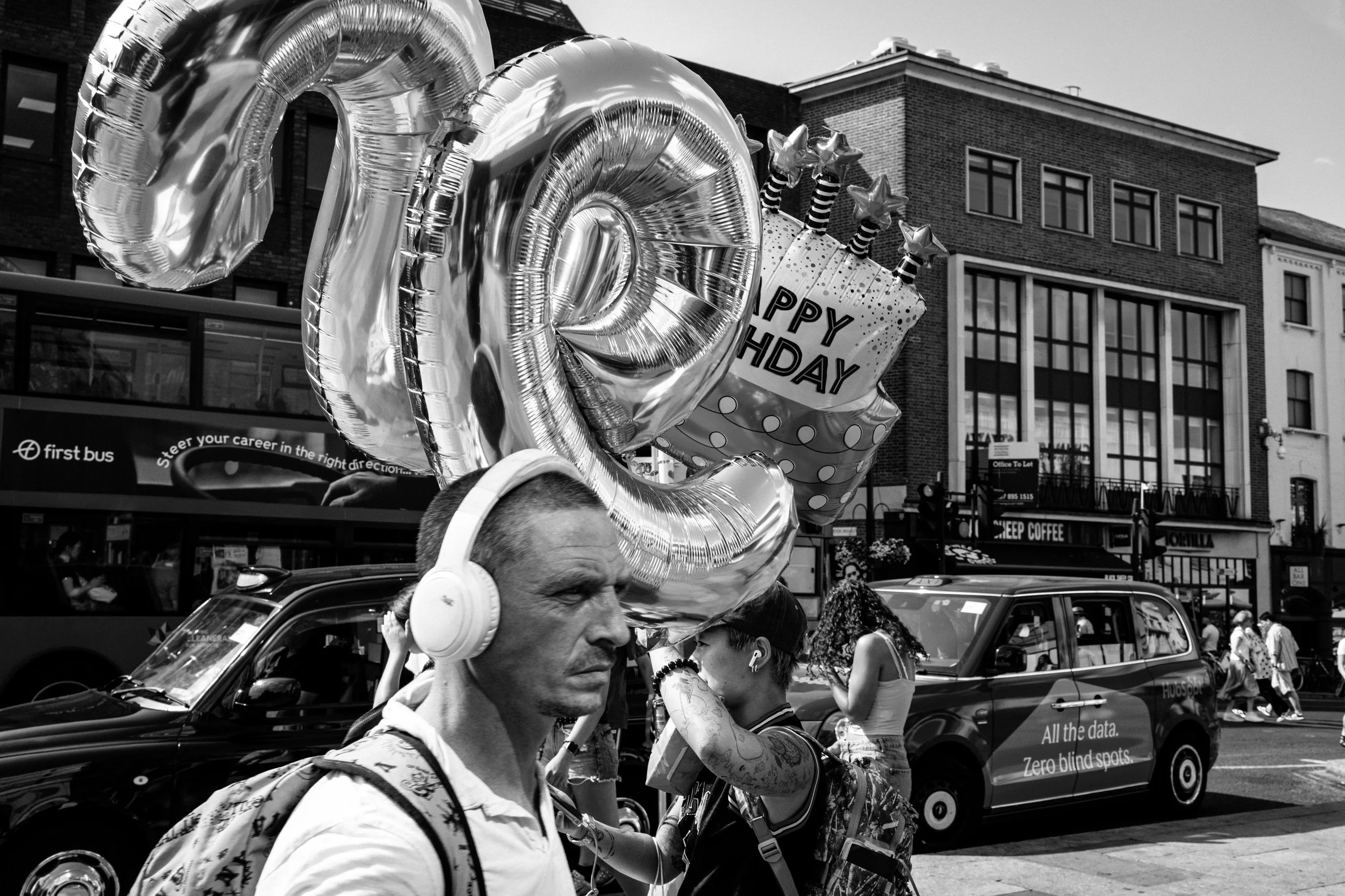 A man with balloons and headphones walking in a busy city street with people and cars around.