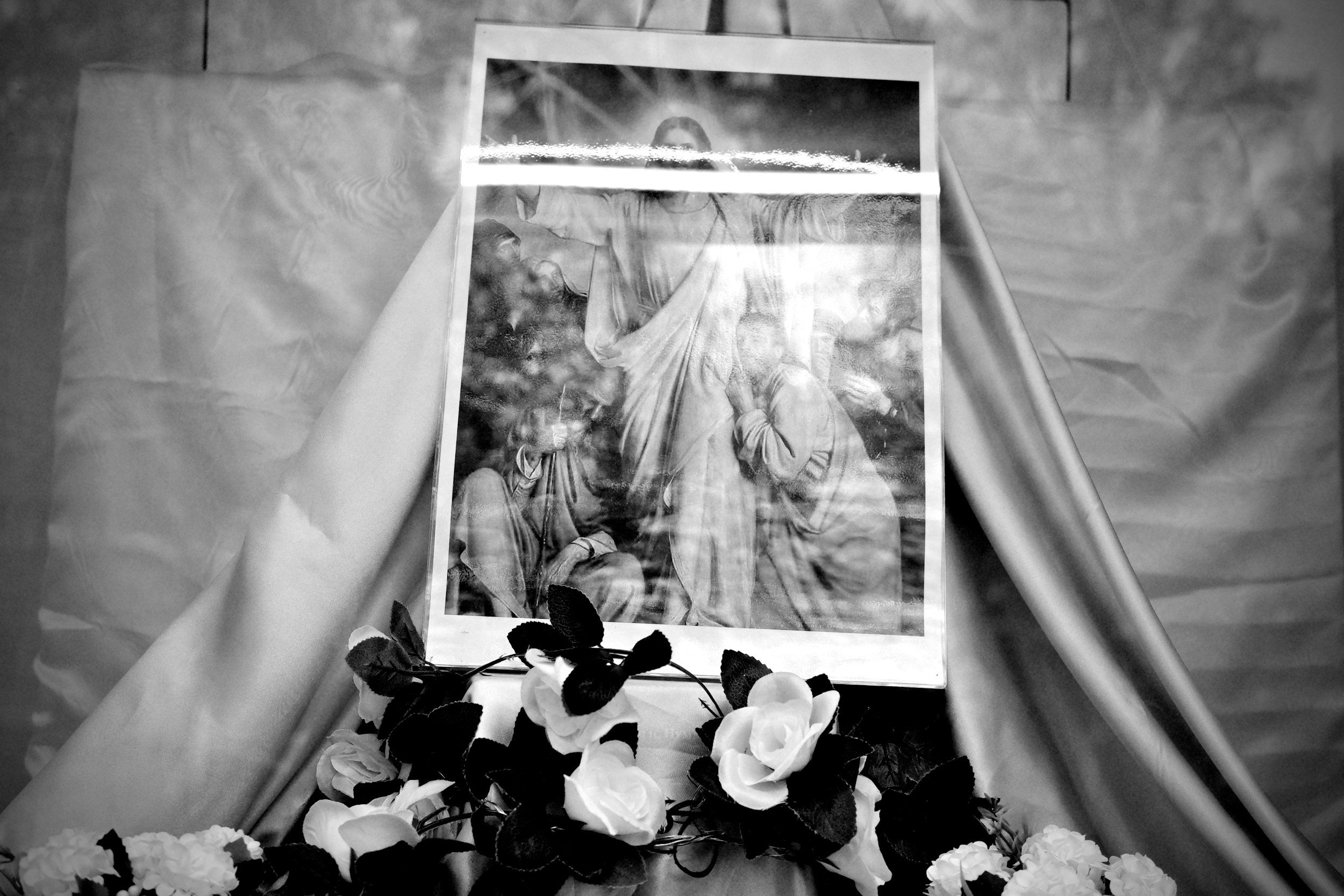 Black and white photograph of a religious shrine with a framed picture of Jesus Christ at the center, surrounded by flowers and candles.