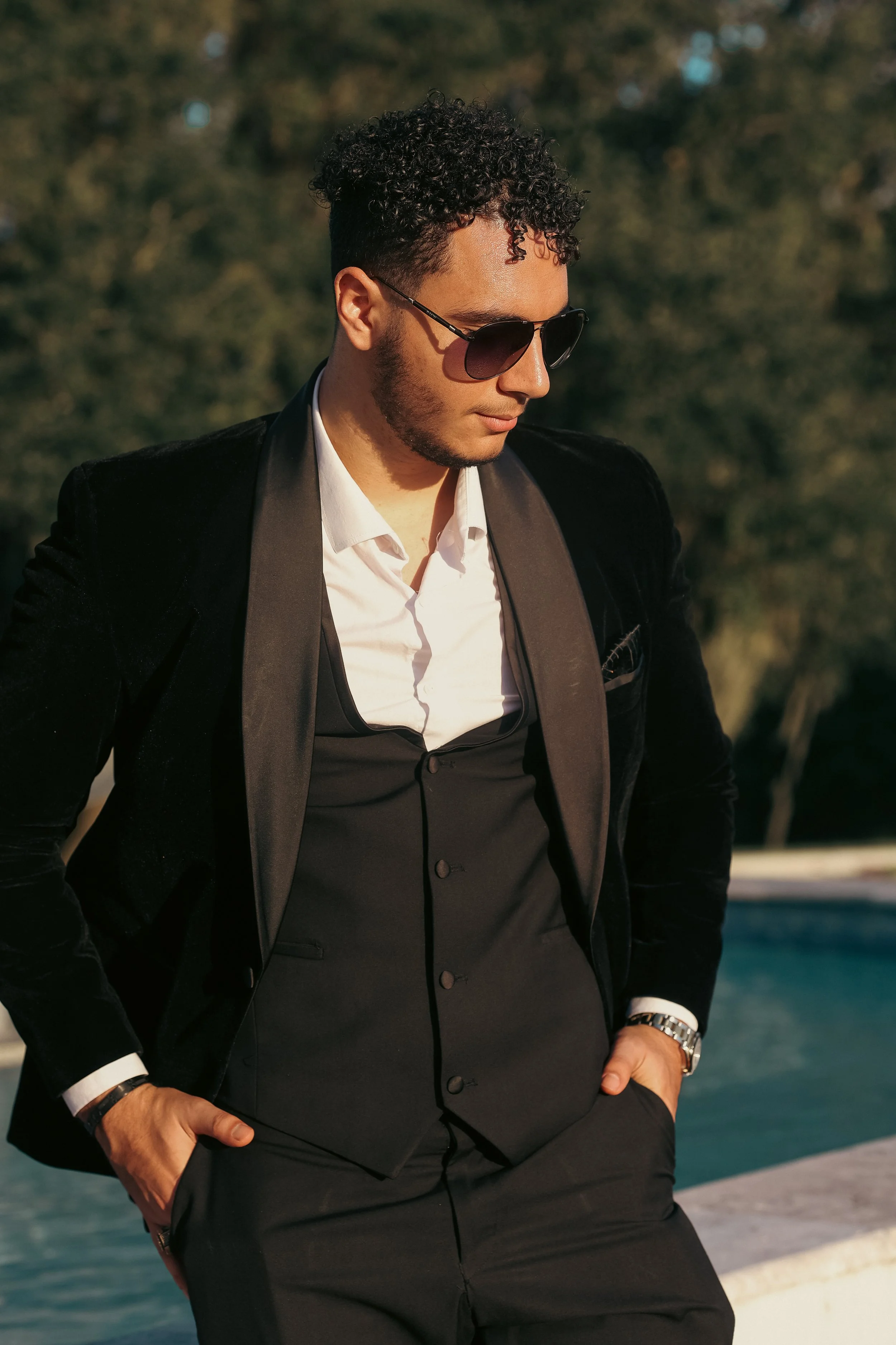 A man in formal attire with sunglasses, wearing a tuxedo with a white shirt and black vest, standing outdoors with trees and water in the background.
