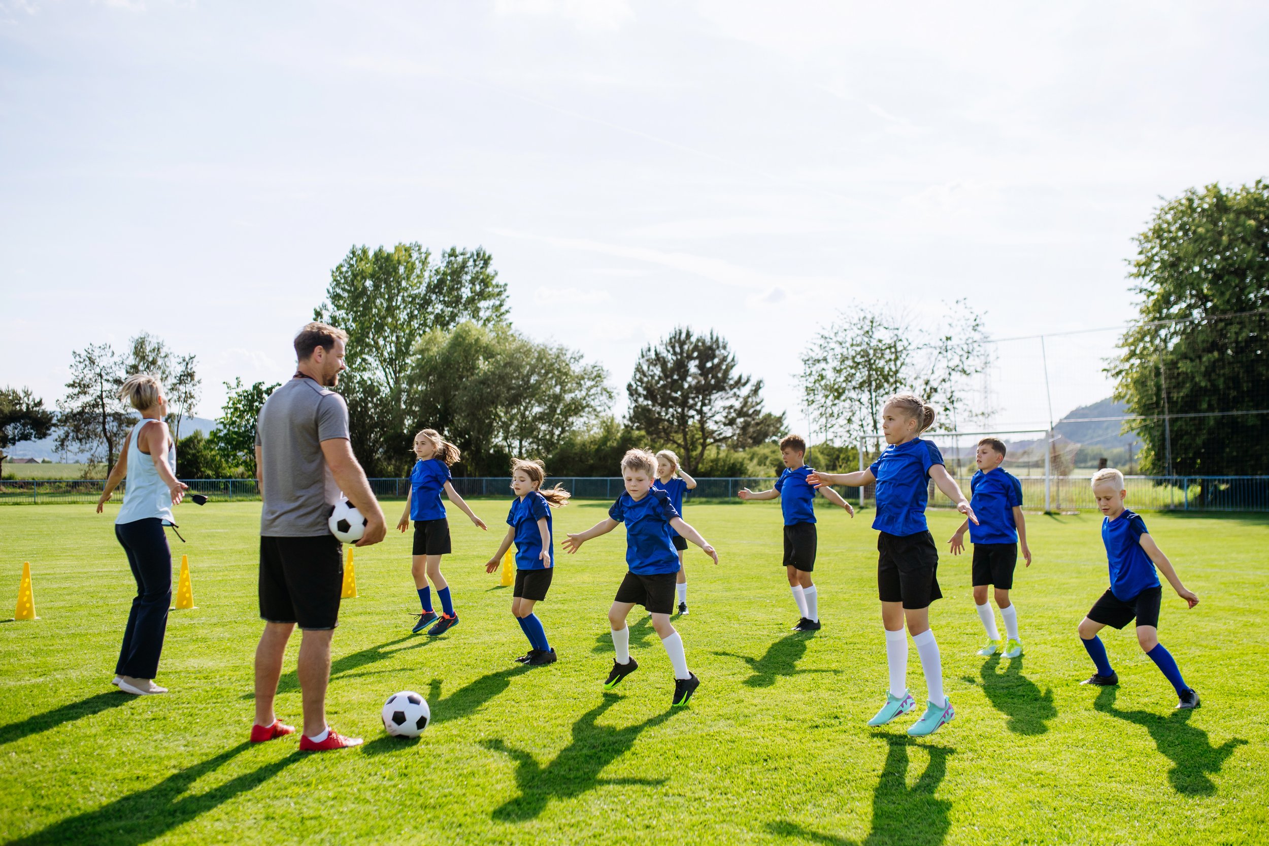 Children and adults participating in a soccer training session on a sunny day. Children are dressed in blue sports uniforms and are stretching or warming up on the grass field, with an instructor or coach guiding them. The field is surrounded by trees and a sports fence.