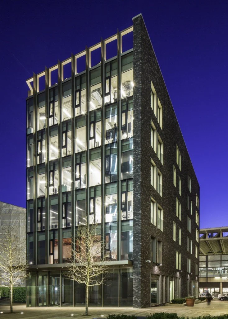 Modern building with multiple stories, large glass windows exposing interior offices, illuminated at night, with trees and sidewalk in the foreground.