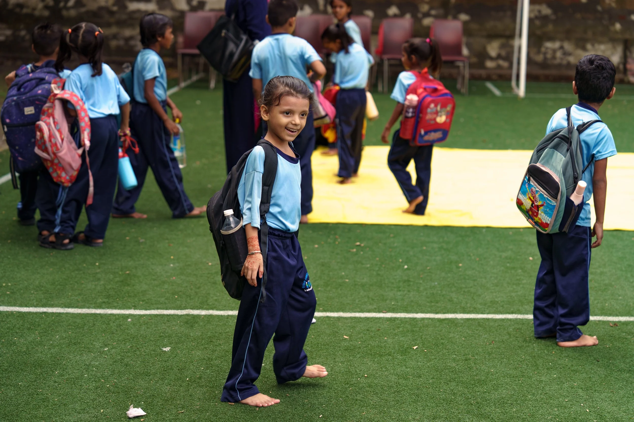 Smiling young girl with a backpack standing on a gym mat outdoors with other children in school uniforms in the background.