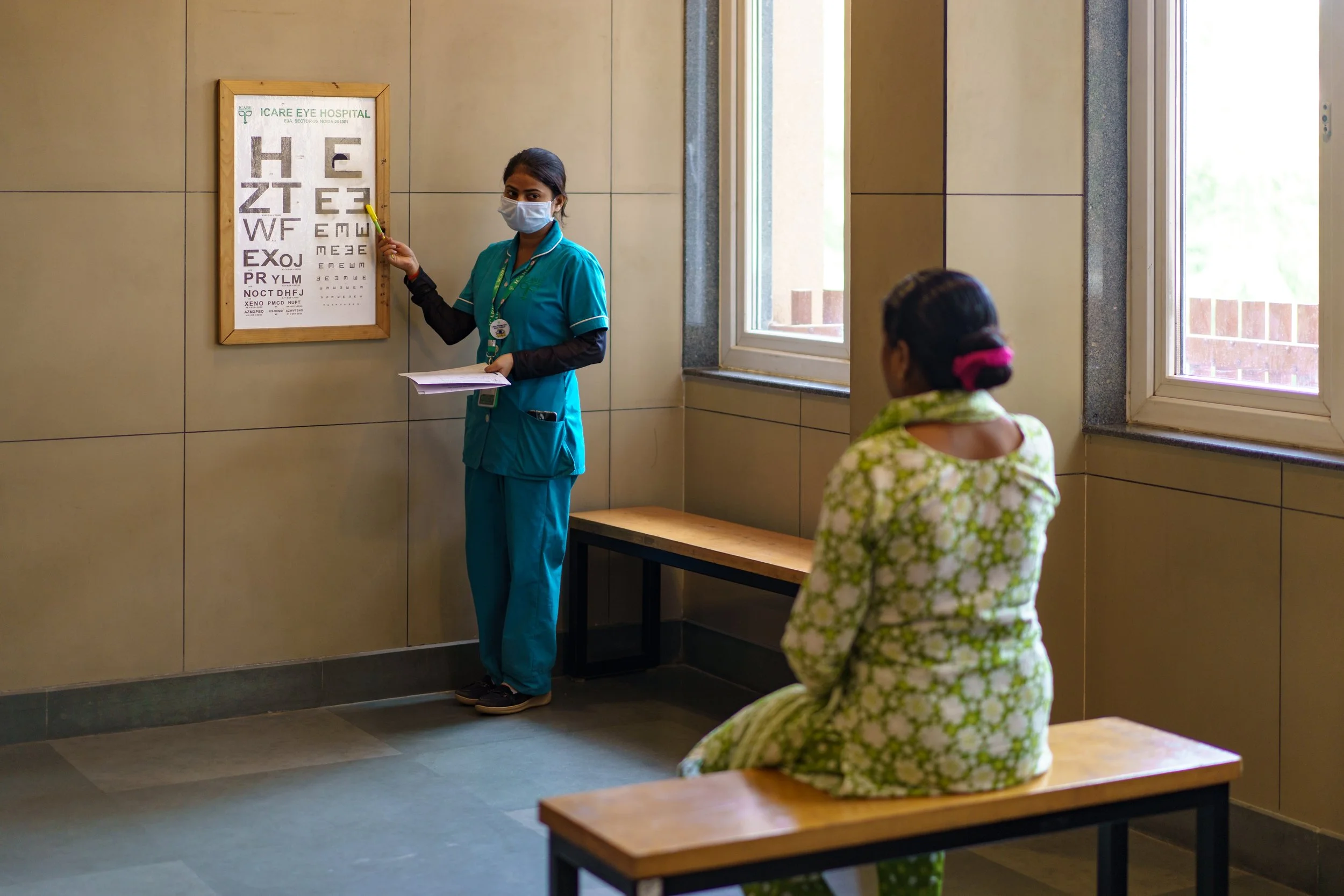 A healthcare professional wearing teal scrubs, a face mask, and a badge points to an eye chart on a wall in an eye hospital. A woman with a pink hair tie and floral dress sits on a bench facing the chart, inside a room with large windows letting in n