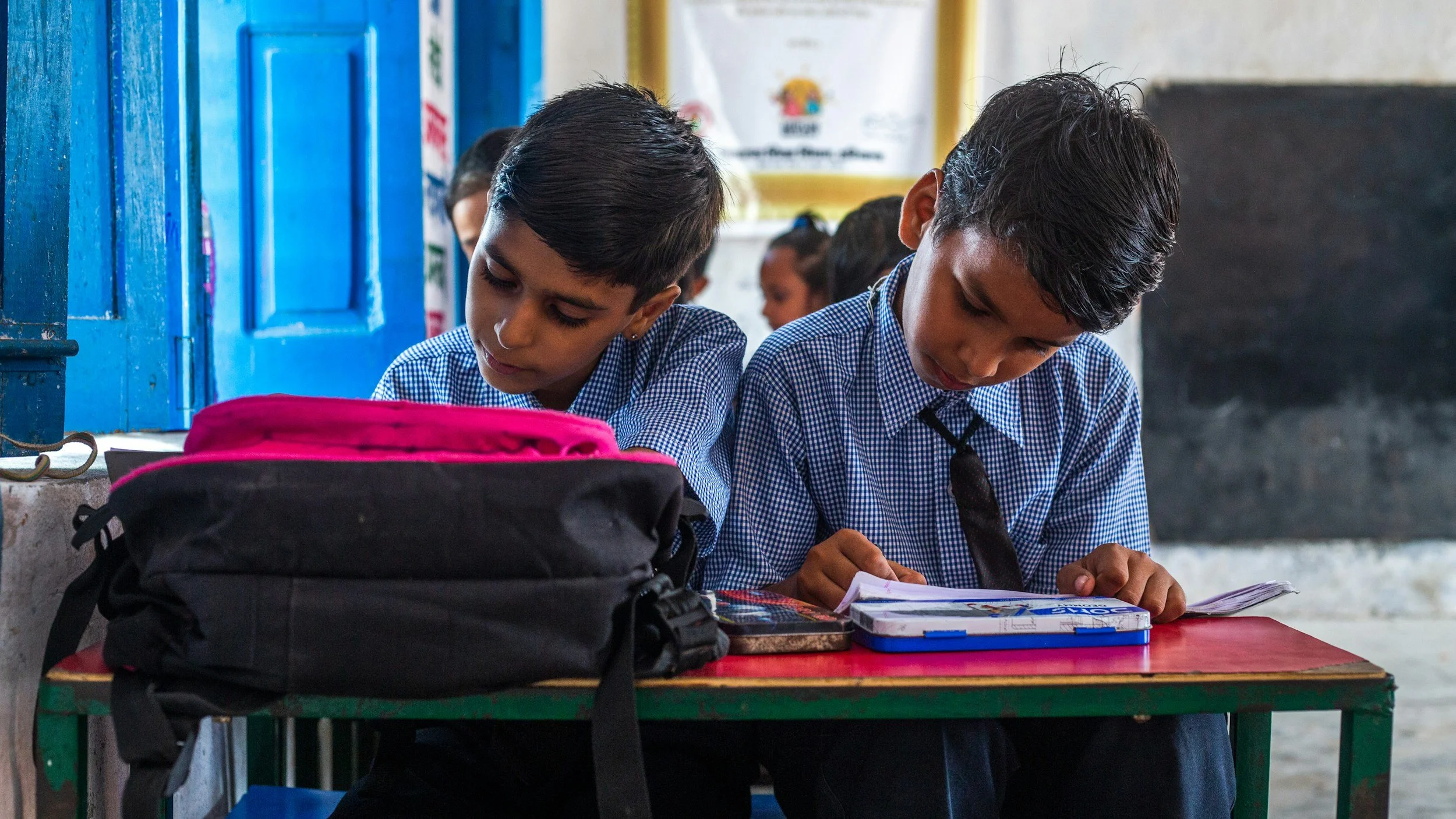 Two young boys in school uniforms sitting at a desk, reading and writing, with notebooks and a pink backpack on the desk. They are in a classroom with blue doors and various posters on the wall.