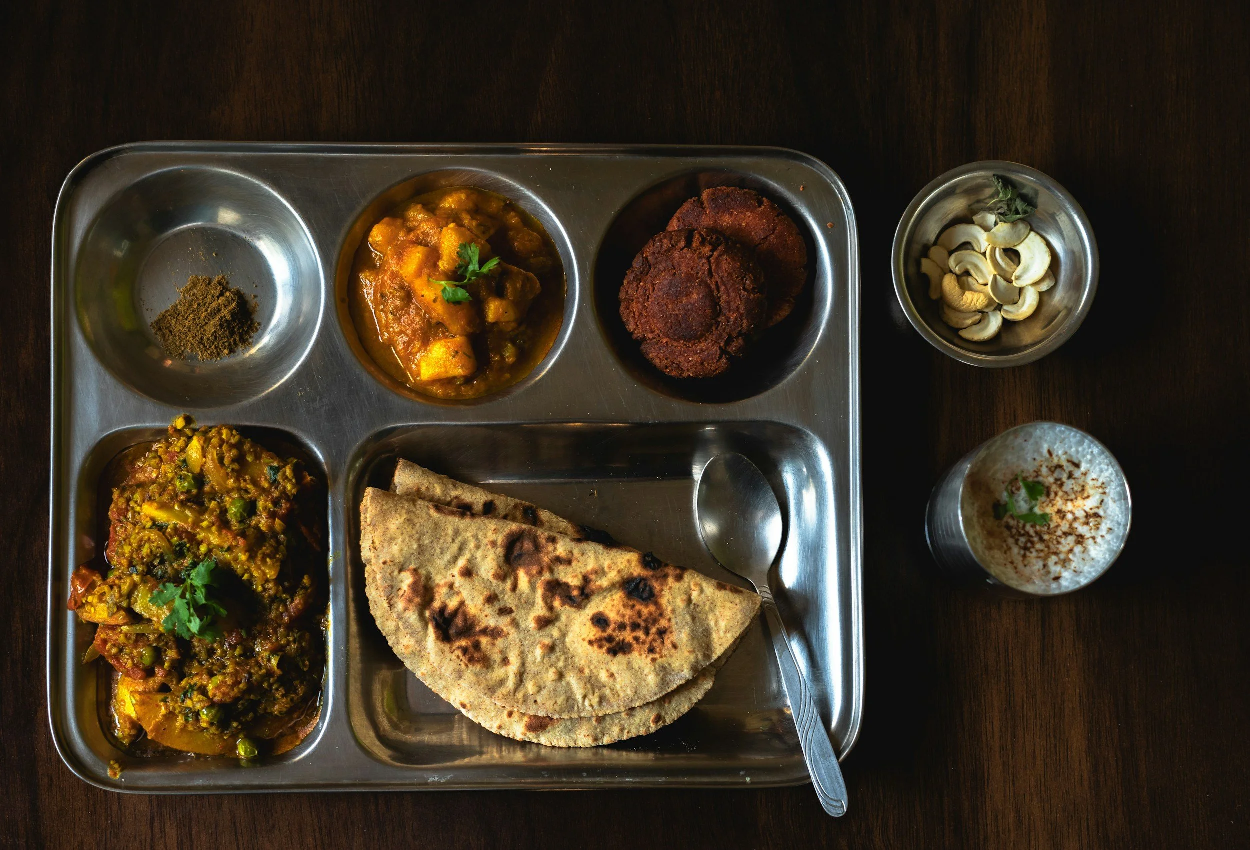 A stainless steel tray with an Indian meal including curry, fried snack, roti, and side dishes, placed on a dark wooden table.