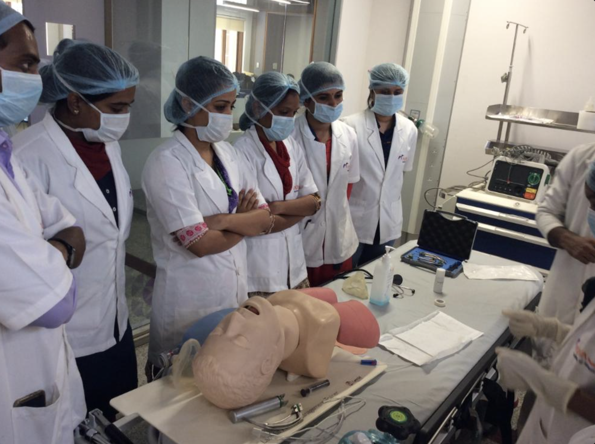 Medical professionals in lab coats, masks, and hair covers gathered around a mannequin and medical equipment in a clinical setting, likely during a training or demonstration.