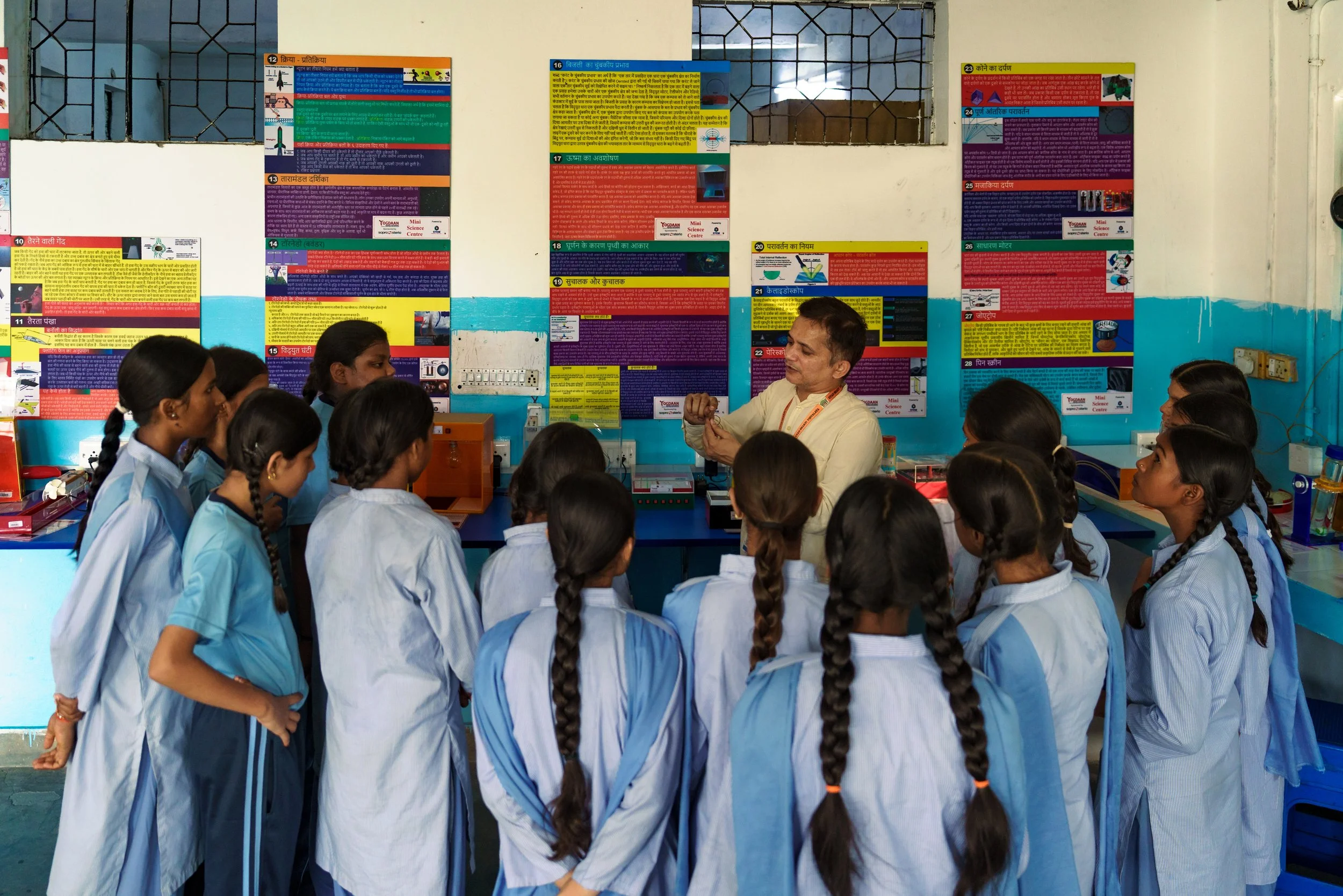 A classroom with a group of students in school uniforms gathered around a teacher who is demonstrating an experiment with a wire and a small device. The classroom walls are decorated with colorful educational posters and charts.