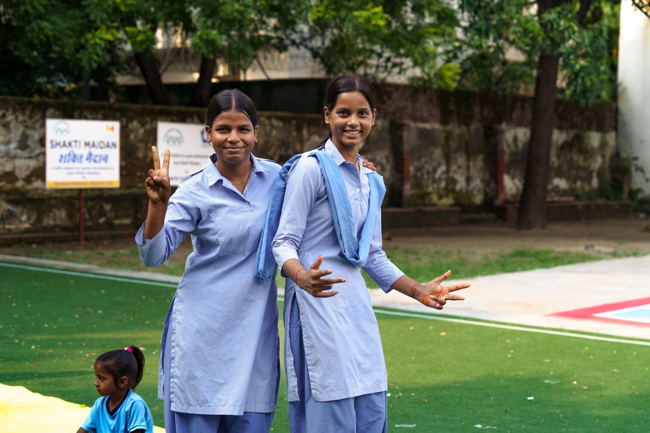 Two young women in light blue school uniforms smiling and making peace signs, walking outdoors on a sports field with trees in the background. A young girl in a blue shirt is sitting on the ground nearby.