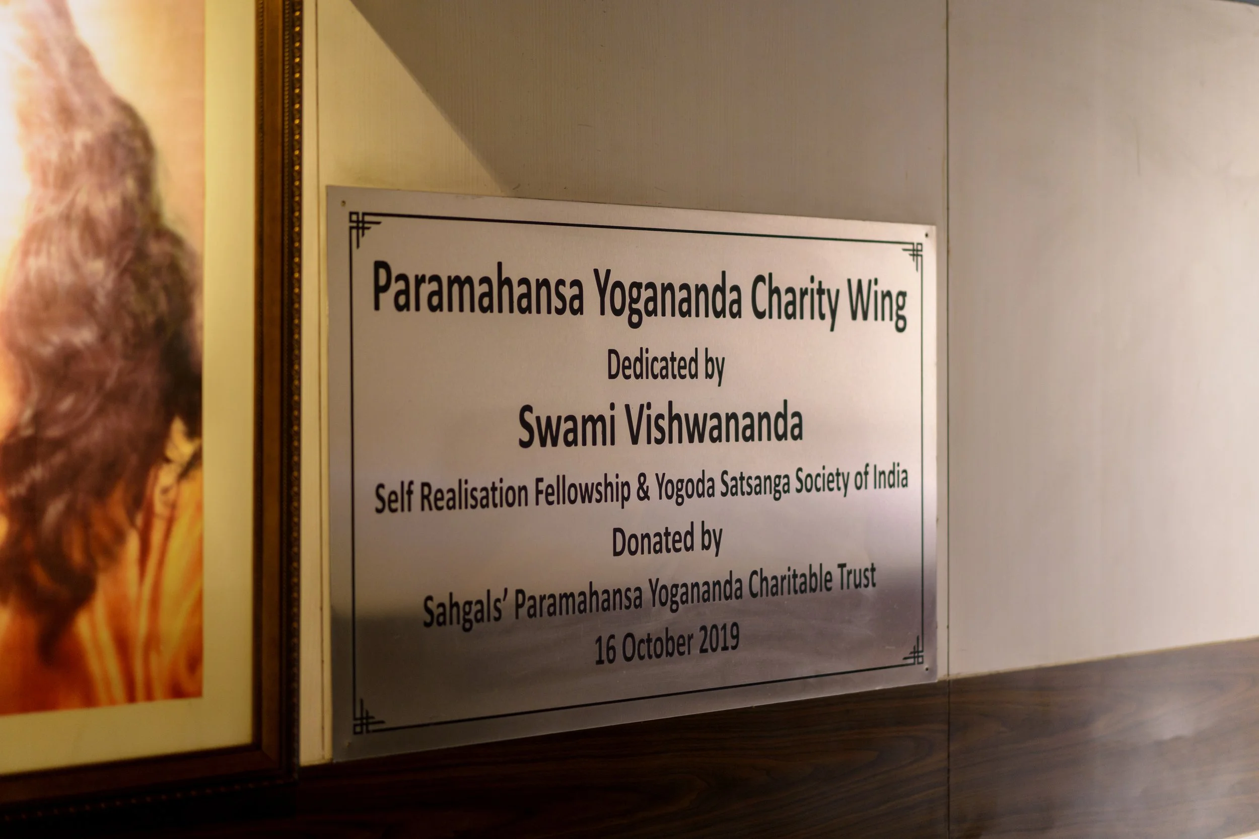  plaque or sign that reads 'Paramahansa Yogananda Charity Wing, Dedicated by Swami Vishwananda, Self Realisation Fellowship & Yogoda Satsanga Society of India, Donated by Swami Vishwananda, Sahgals Paramahansa Yogananda Charitable Trust' along with a