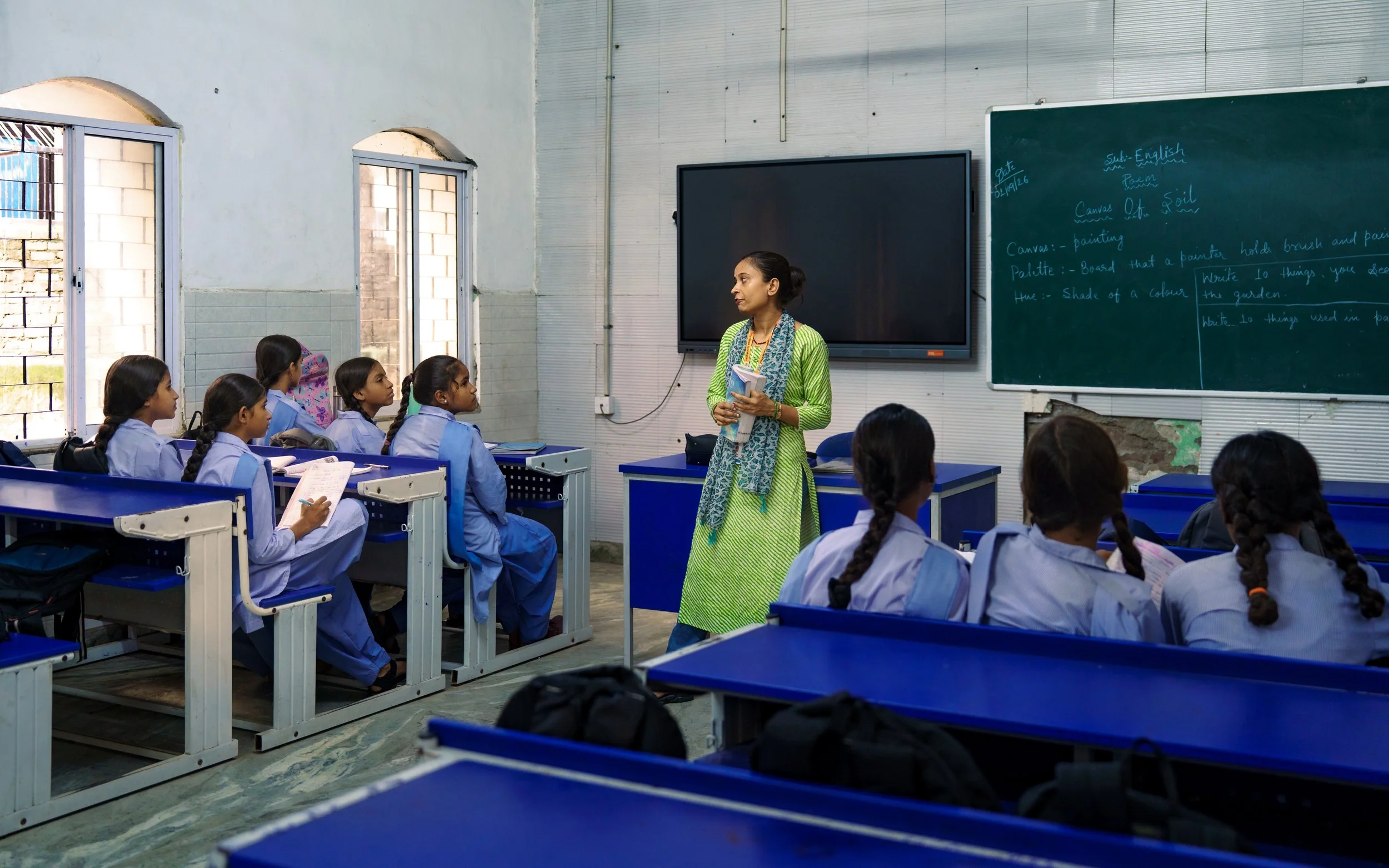 A classroom with a group of young female students in blue uniforms seated at blue desks, attentively listening to a female teacher who is standing near a blackboard and a large digital display. The classroom has open windows with sunlight coming in.