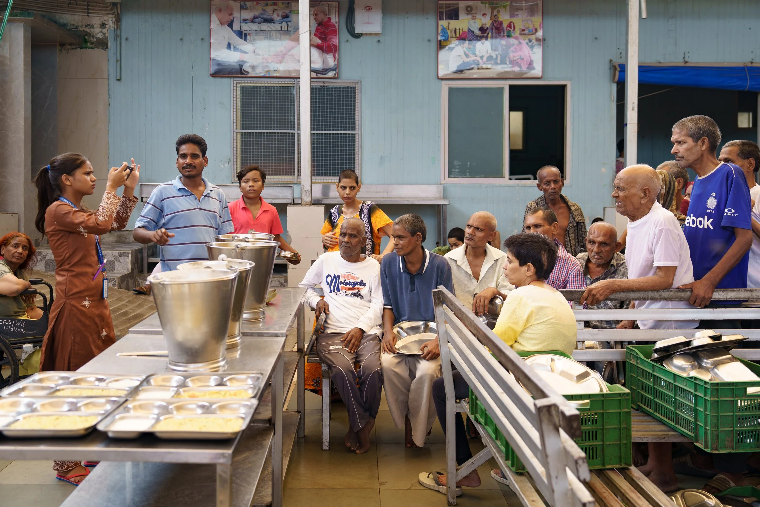 A crowded cafeteria with people waiting in line and seated, during meal service, with various food containers and trays.