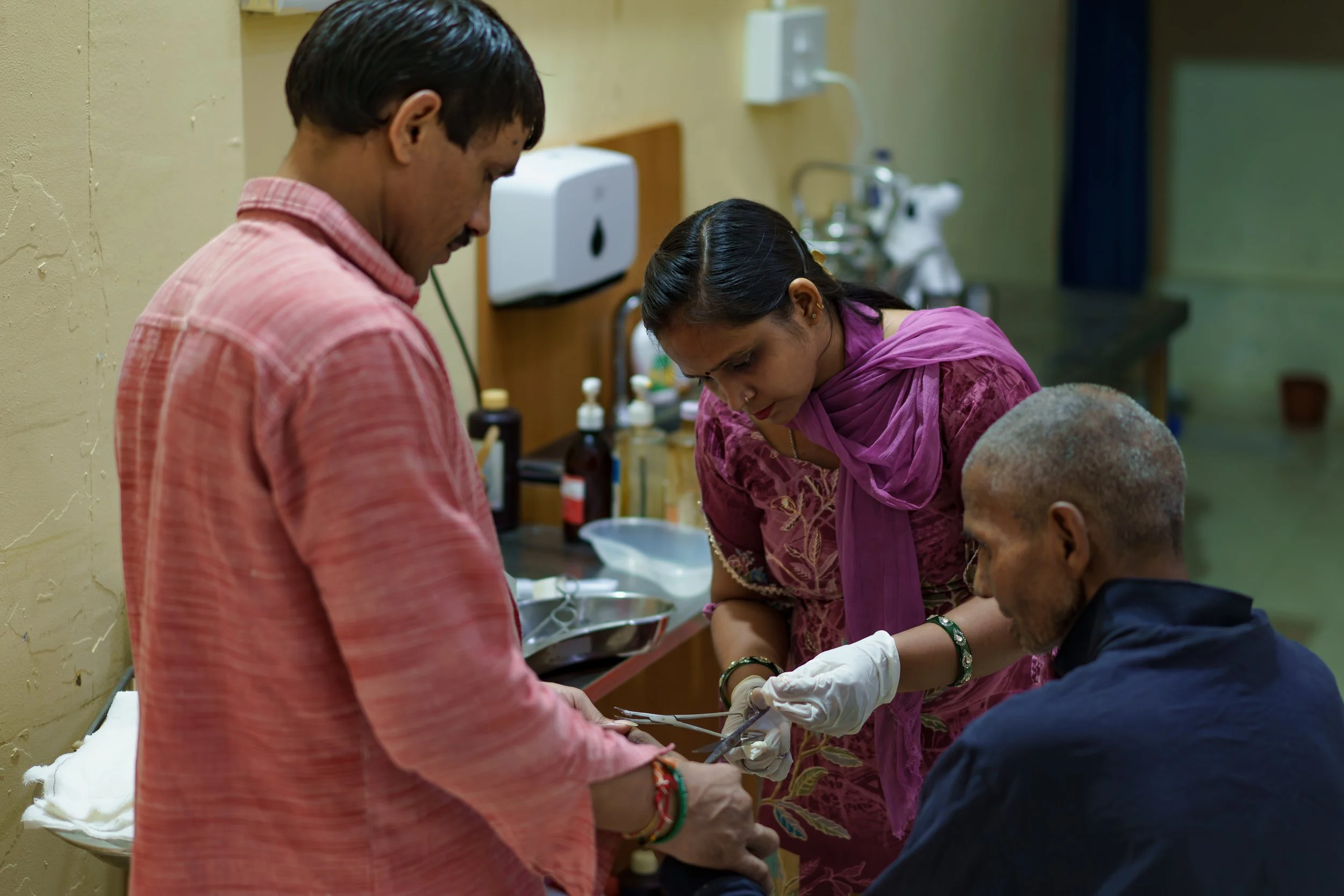 A group of four people in a clinical setting, with one man wearing a pink shirt, a woman in traditional Indian attire, and two other individuals. They are engaged in a healthcare procedure, with some wearing gloves and focused on a task.