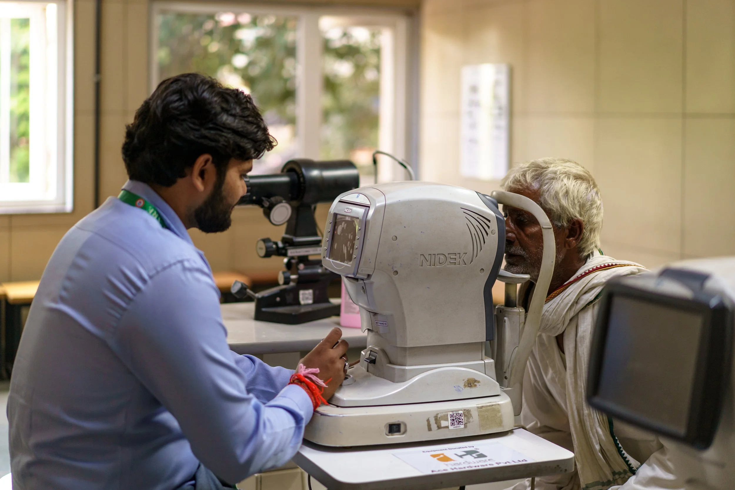 An eye exam being conducted on an elderly man by a healthcare professional in a clinic. The man is sitting in front of a machine used for eye testing, while the professional is adjusting the equipment.