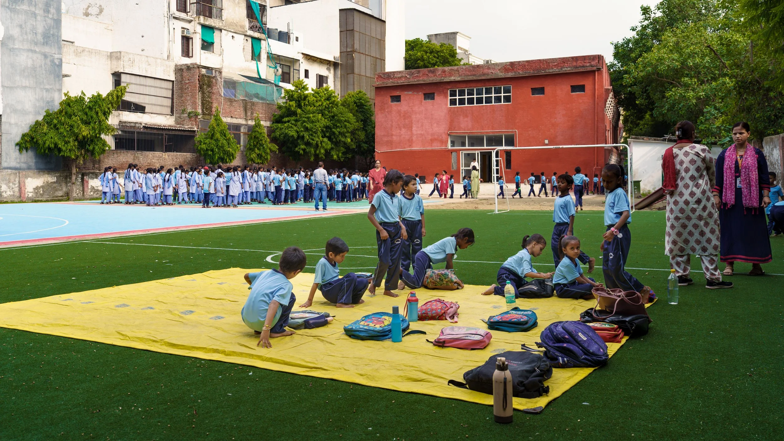 Students and teachers gather in a schoolyard for an outdoor activity. Children are seated on a yellow mat and standing in lines on the green lawn, with backpacks and water bottles nearby. Some students are wearing light blue uniforms. Teachers are su