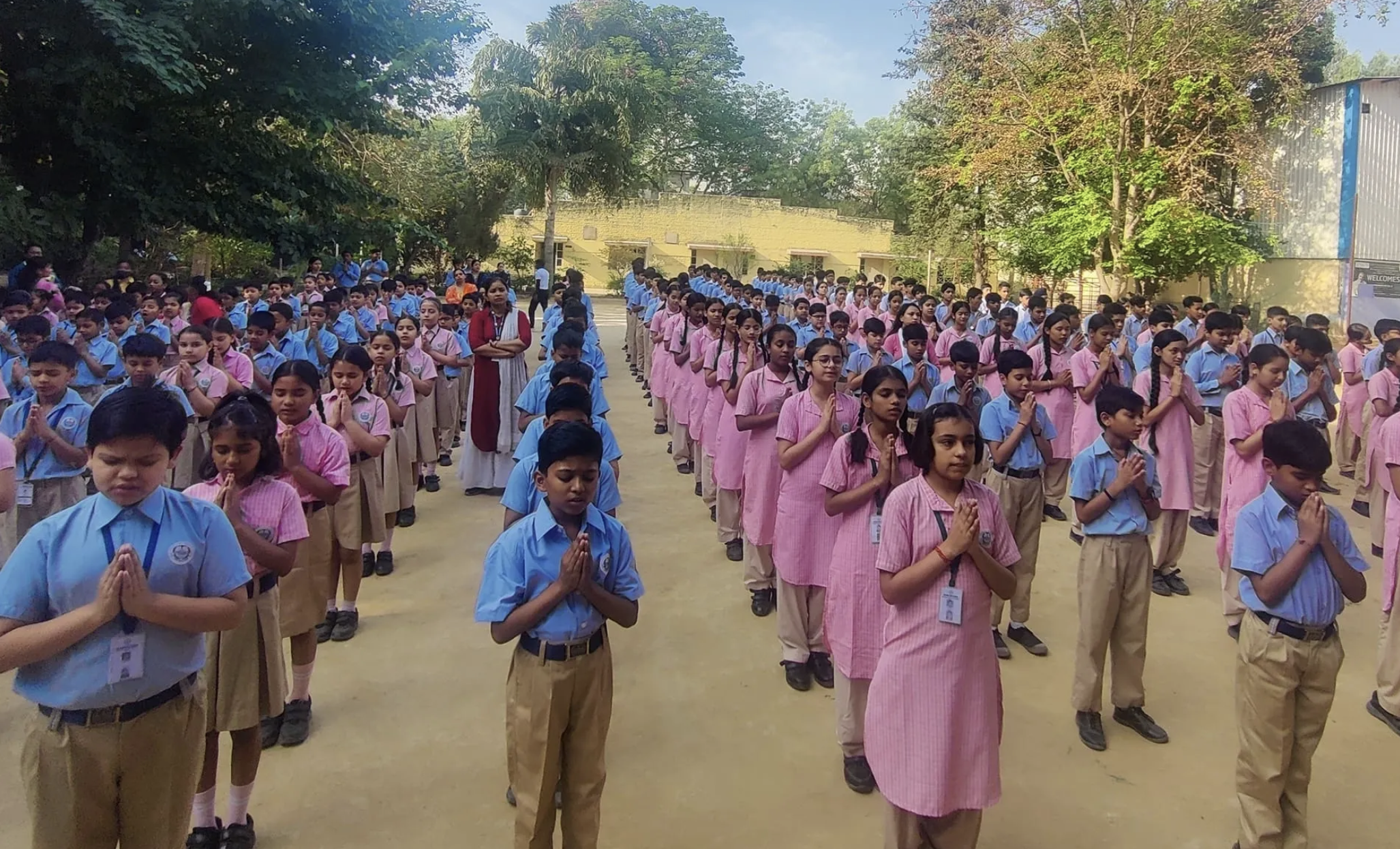 School children standing outdoors, practicing prayer with hands pressed together in a respectful gesture, wearing uniforms in blue and pink, accompanied by teachers, surrounded by trees.