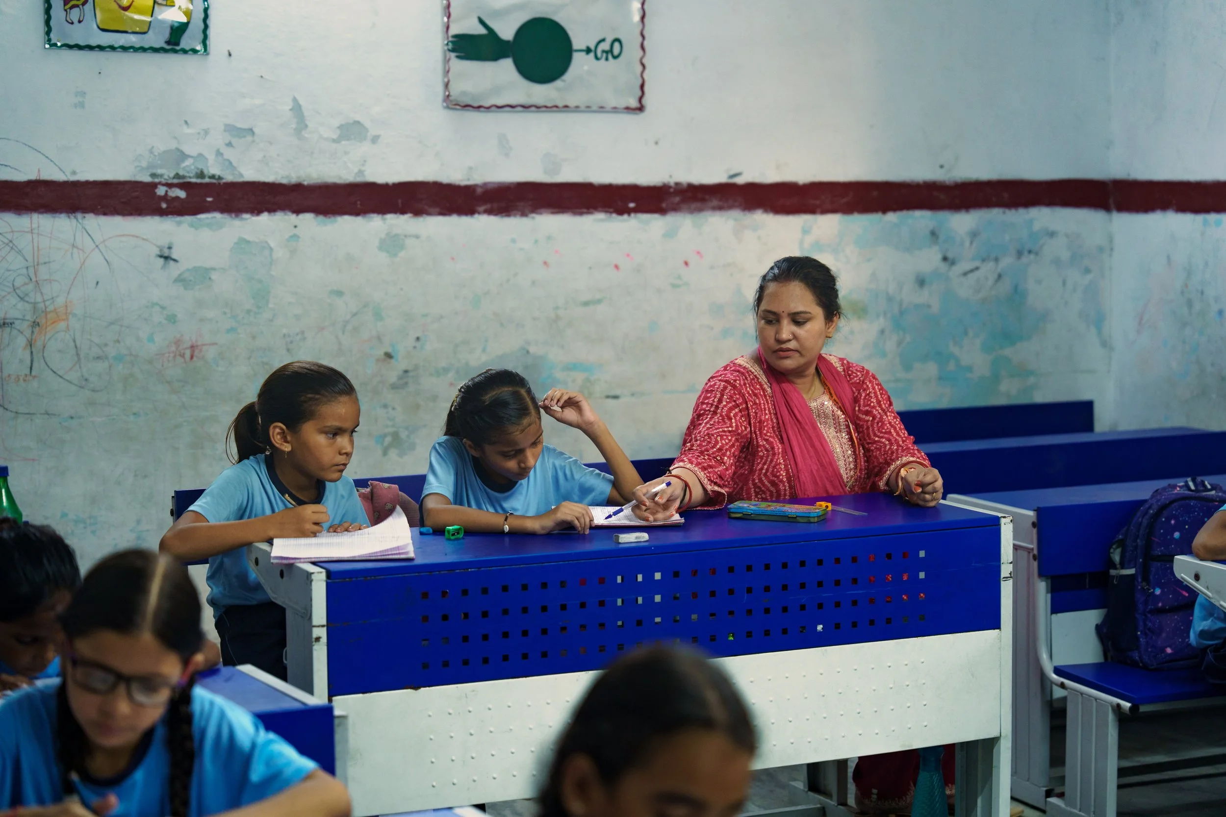 A classroom scene with a female teacher in a red patterned sari overseeing two young students in blue uniforms working at a blue desk. Several students are visible in the foreground. The background shows a wall with some posters and peeling paint.