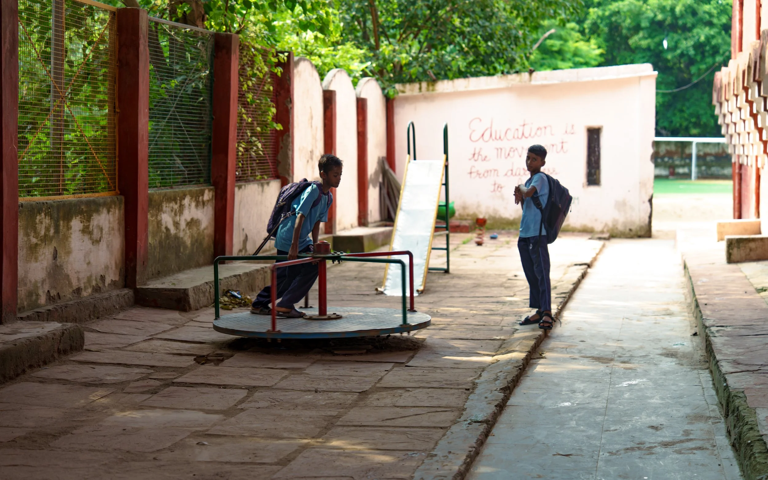 Two children with backpacks are near a merry-go-round in an outdoor area, with a white wall that has red text reading 'Education is the movement from darkness to light' and lush green foliage in the background.