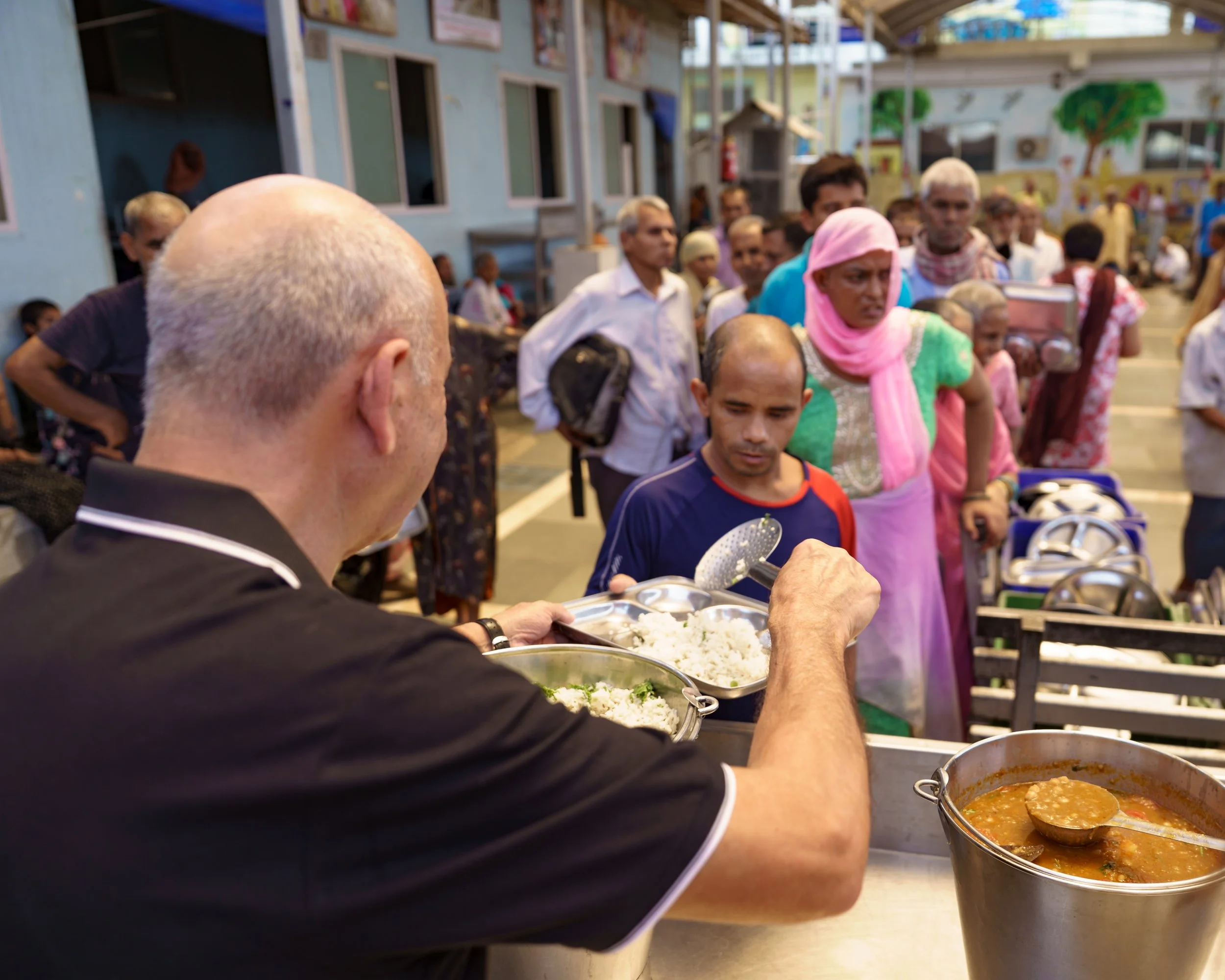 A man serving rice and curry at a food distribution event, with a large crowd waiting in line under a covered outdoor area.