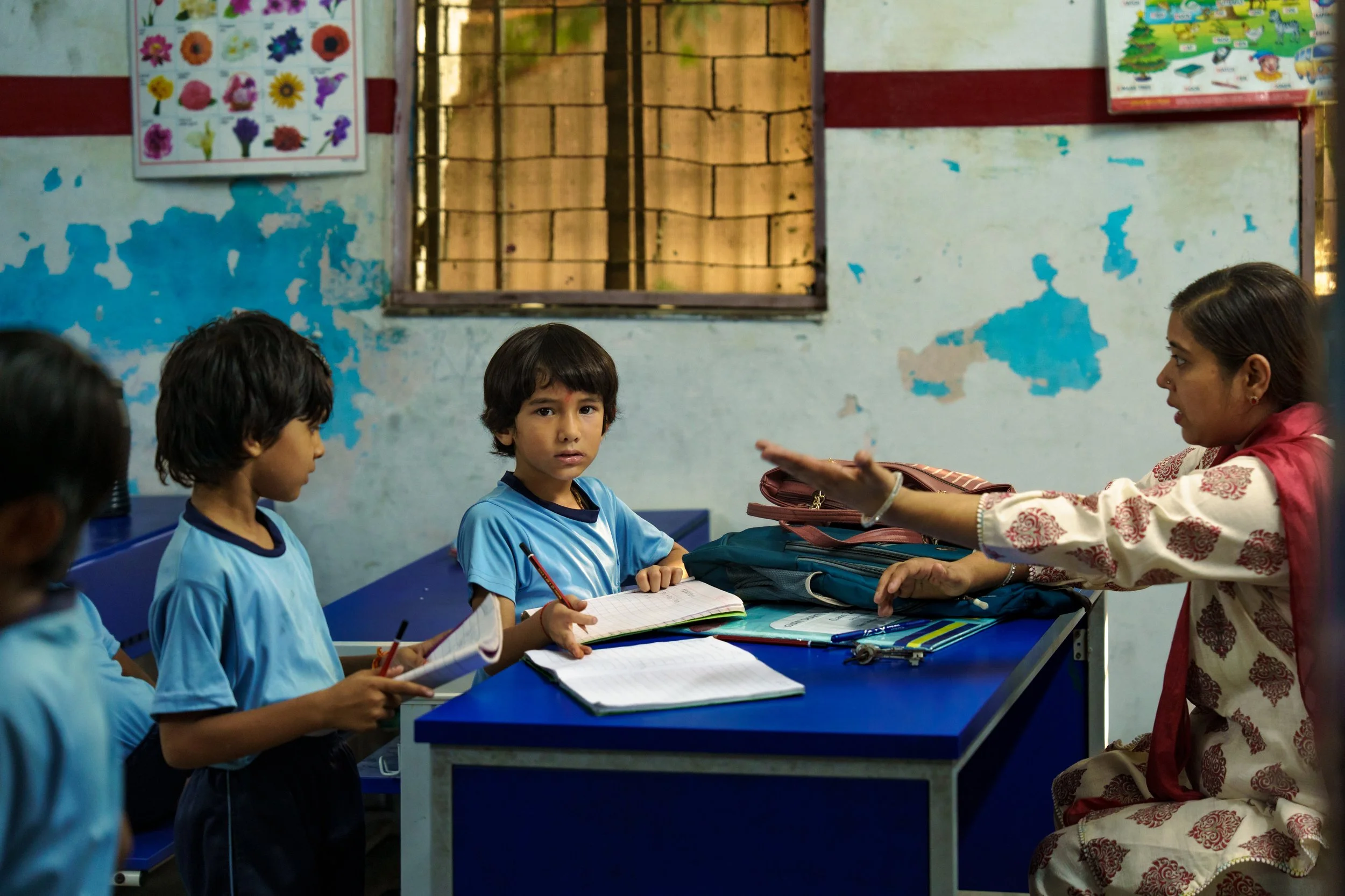 A classroom scene with a woman teacher talking to students wearing blue uniforms, sitting at desks, while holding notebooks and pens. The classroom has weathered walls with blue paint patches and educational posters on the walls.
