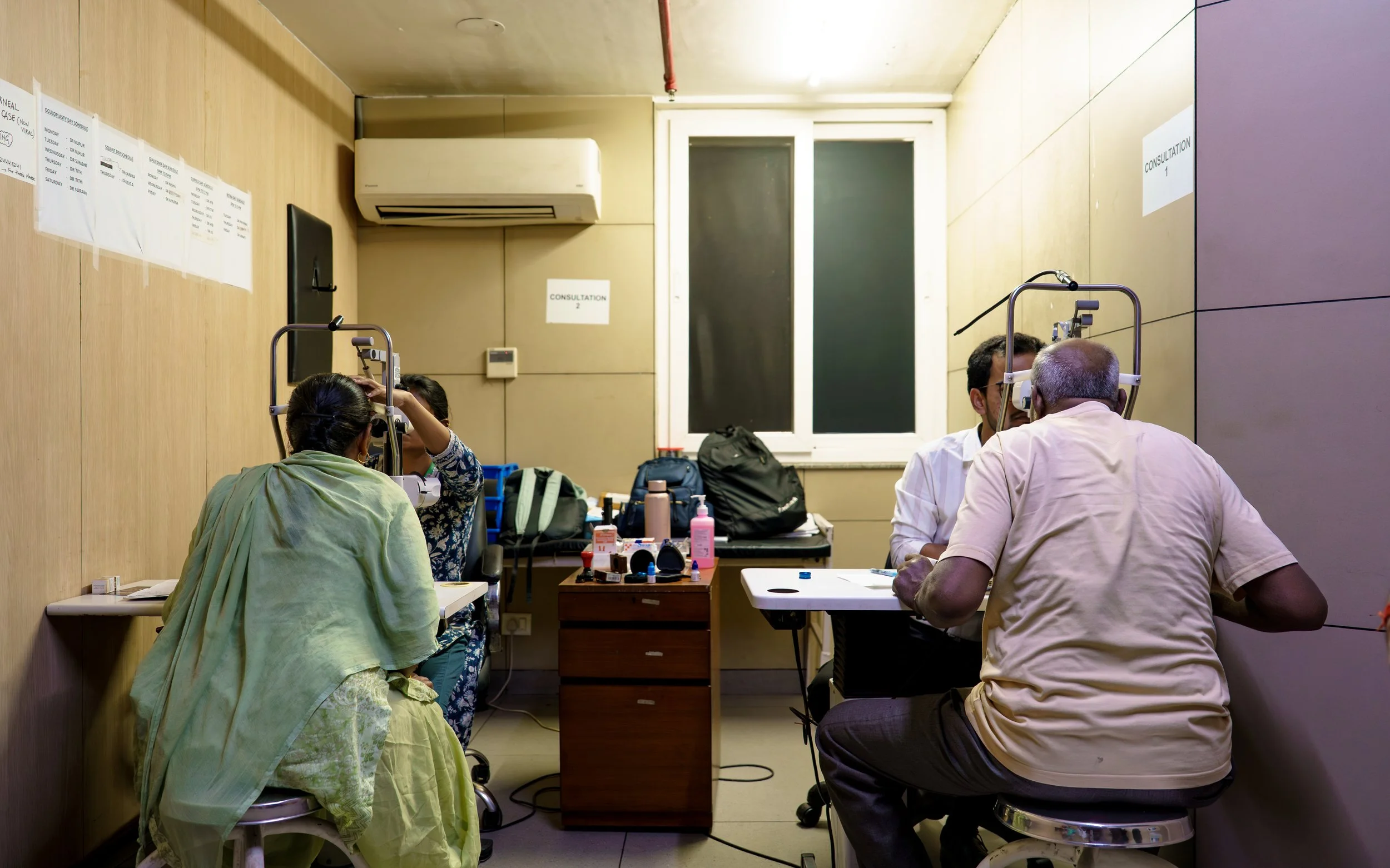 People getting eye examinations in a clinic, with equipment and supplies on a table, and signs on the walls indicating consultation areas.