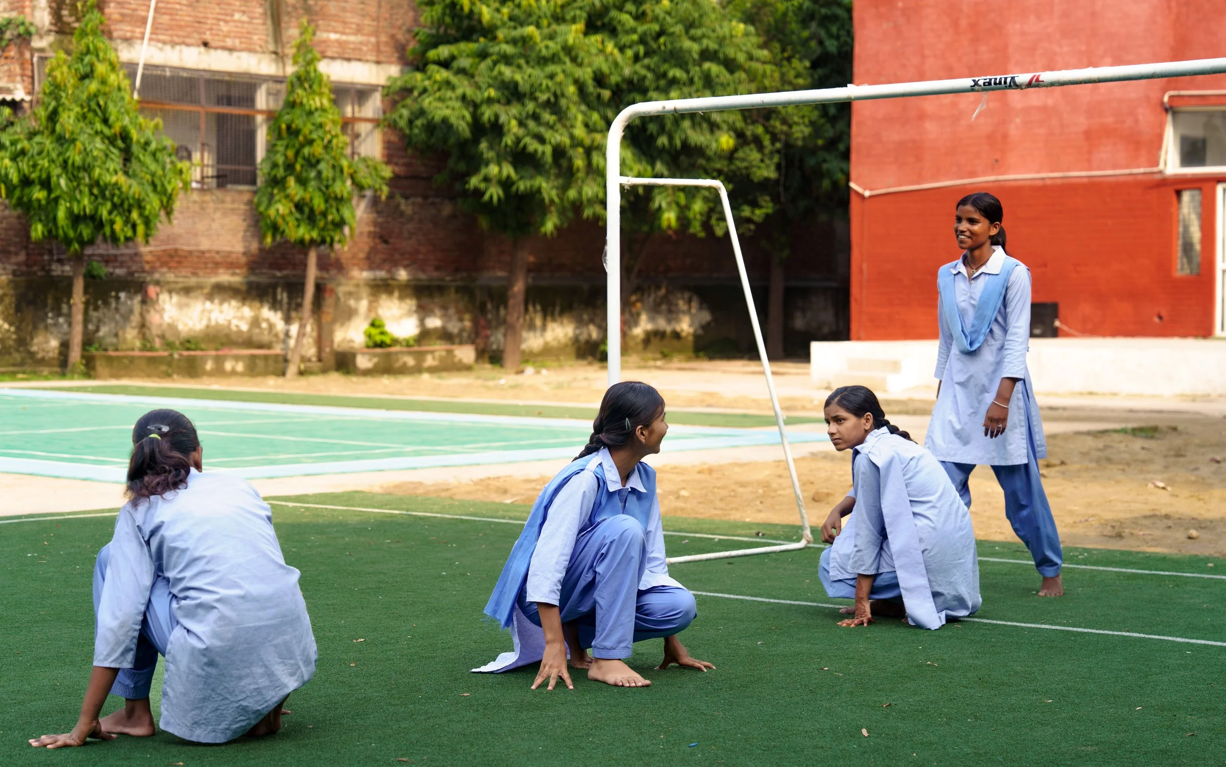 A group of young girls wearing blue uniforms are practicing yoga or a similar activity outdoors on a sports field with a goalpost nearby, surrounded by trees and brick buildings.