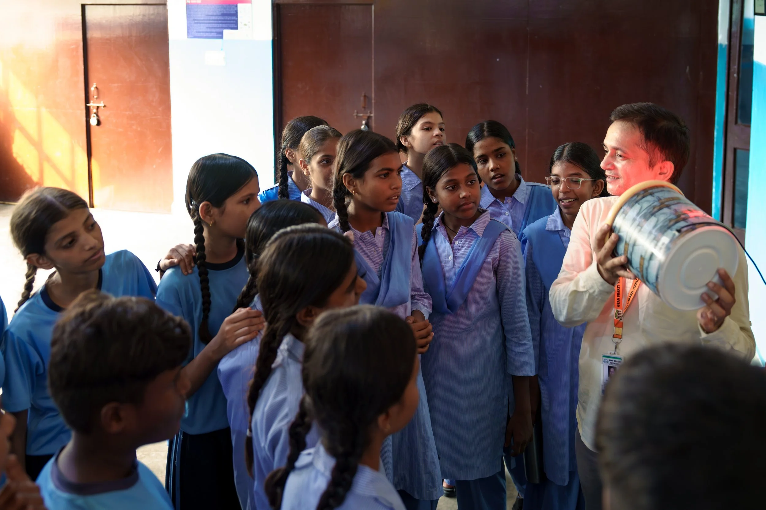 A group of schoolgirls, dressed in blue uniforms, listens to a man holding a large globe-shaped object.