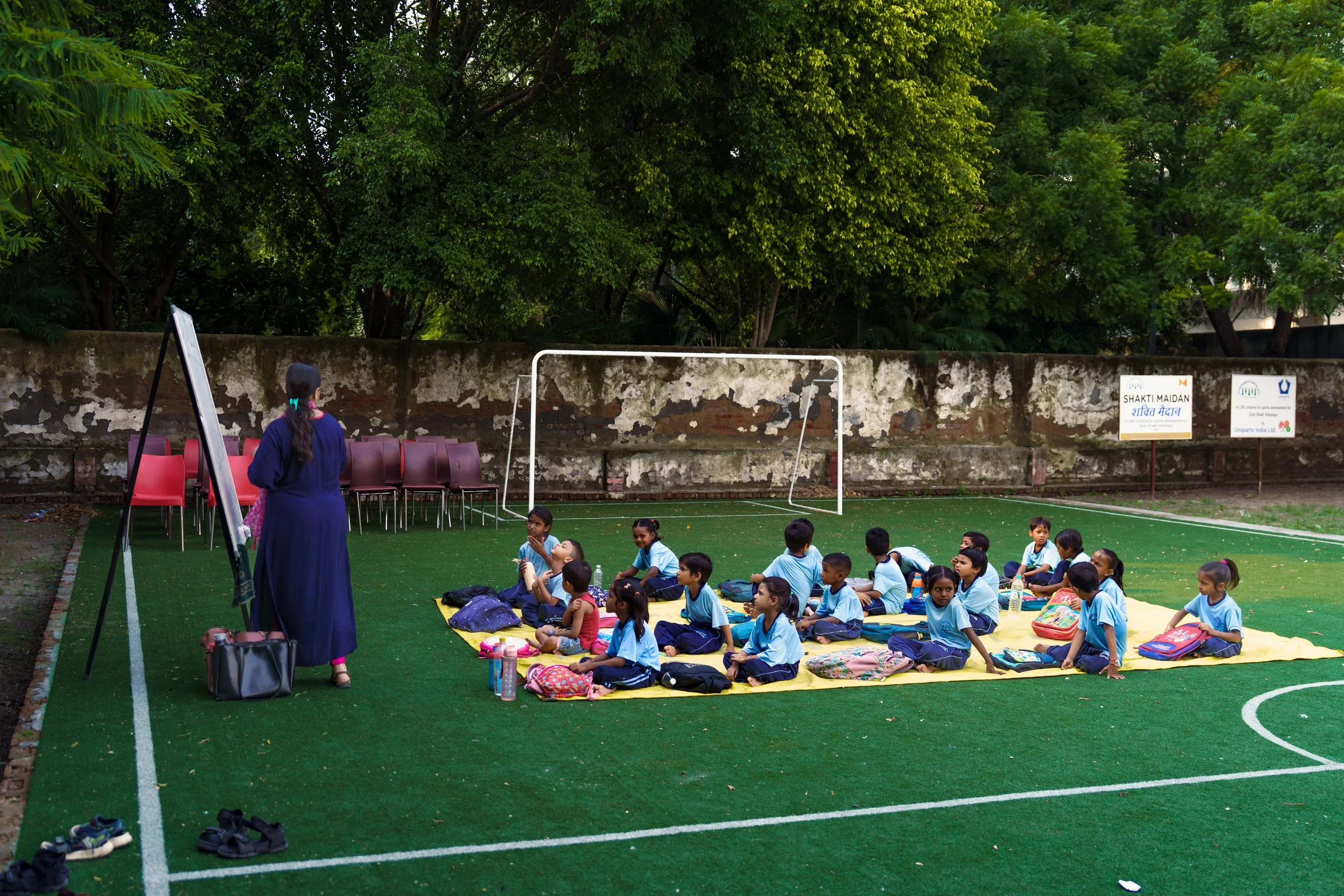 Children sitting on a yellow mat outdoors, listening to a woman in traditional Indian attire, with a soccer goal, chairs, and a wall with trees in the background.