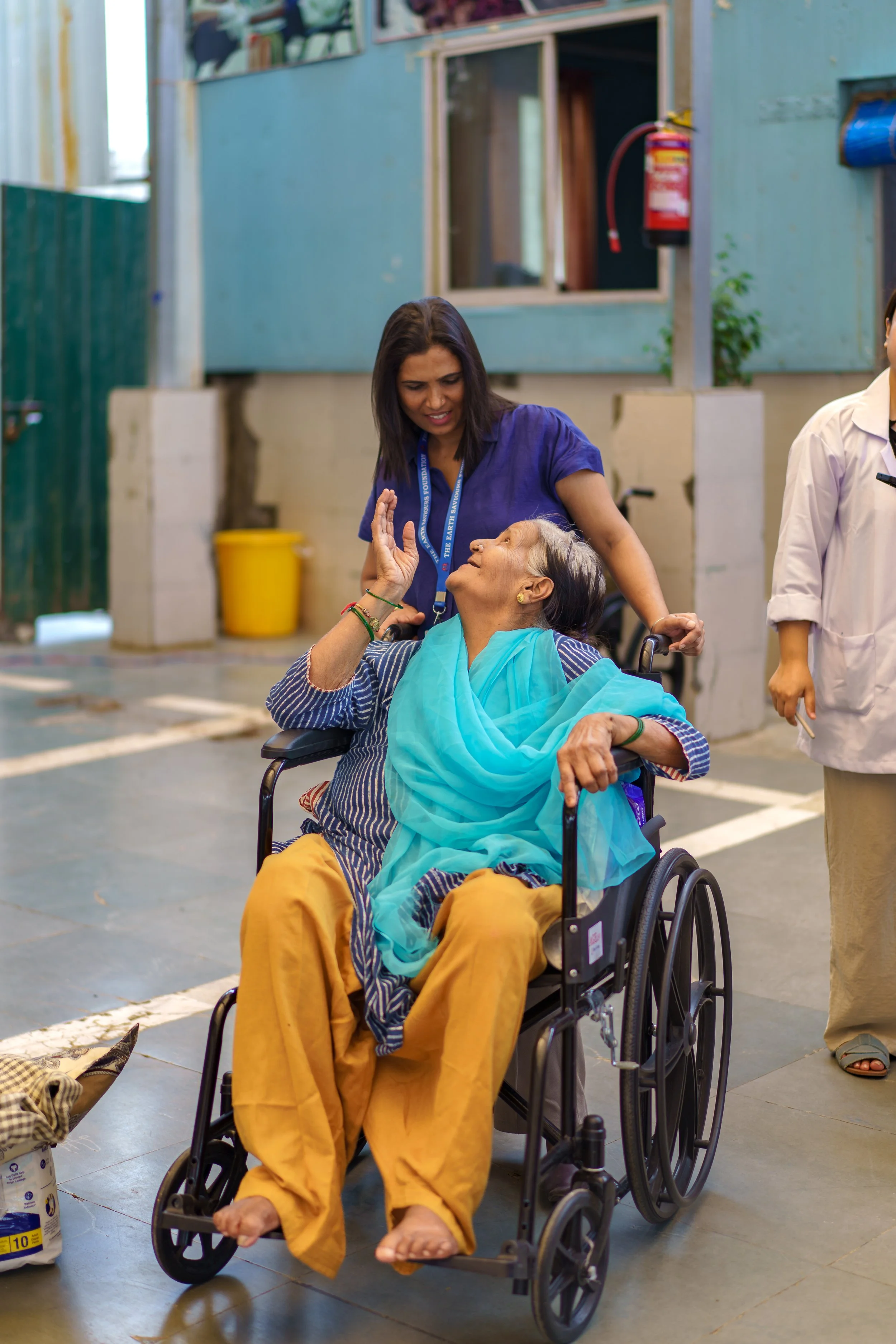 A woman in a wheelchair reaches out to touch the face of an elderly woman sitting in the wheelchair, both smiling and engaging with each other indoors.
