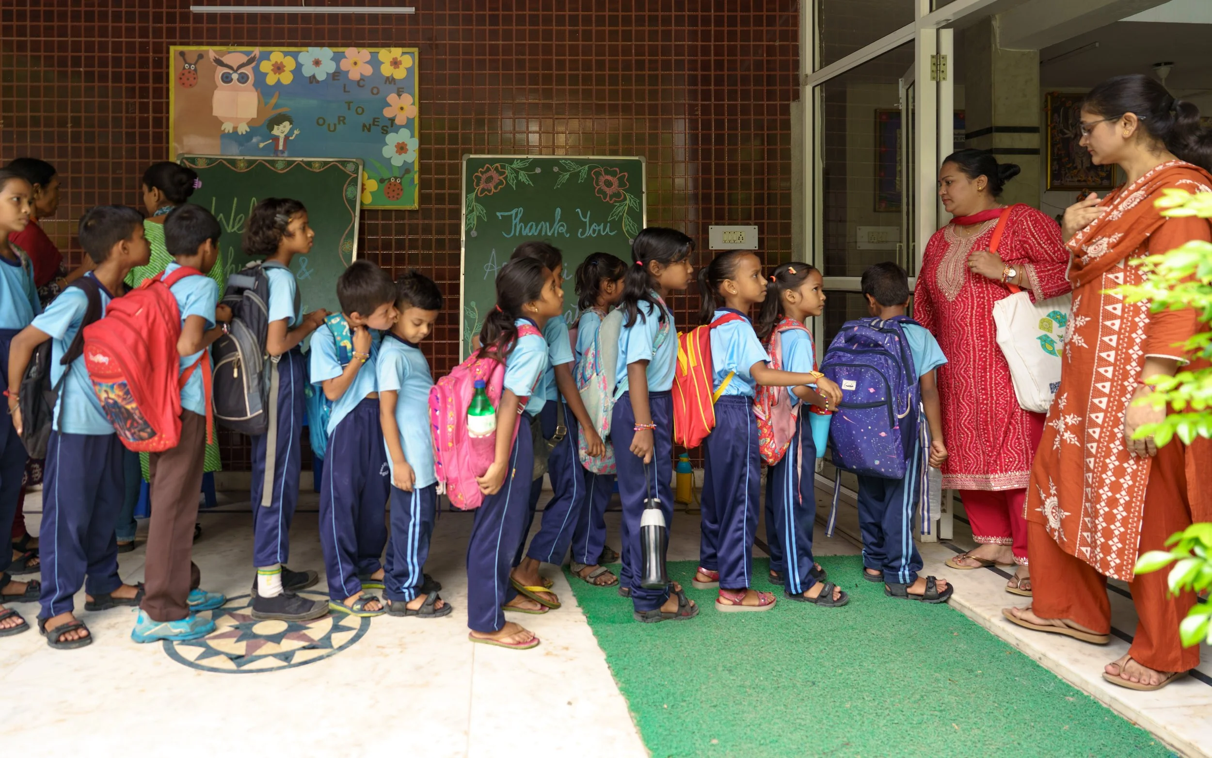 A line of young children wearing blue uniforms and backpacks waiting outside a school entrance as two women, dressed in traditional attire, speak to them.