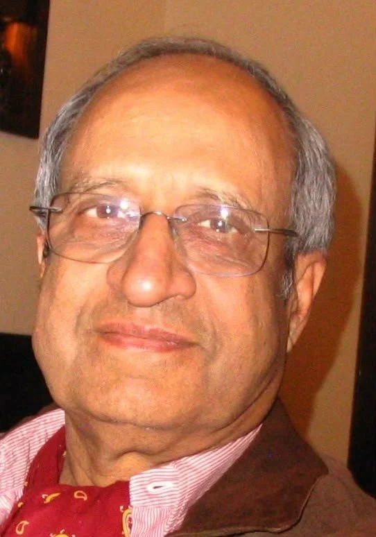 A close-up photo of an older man with gray hair, wearing glasses, a red necktie, and a brown jacket, smiling slightly.