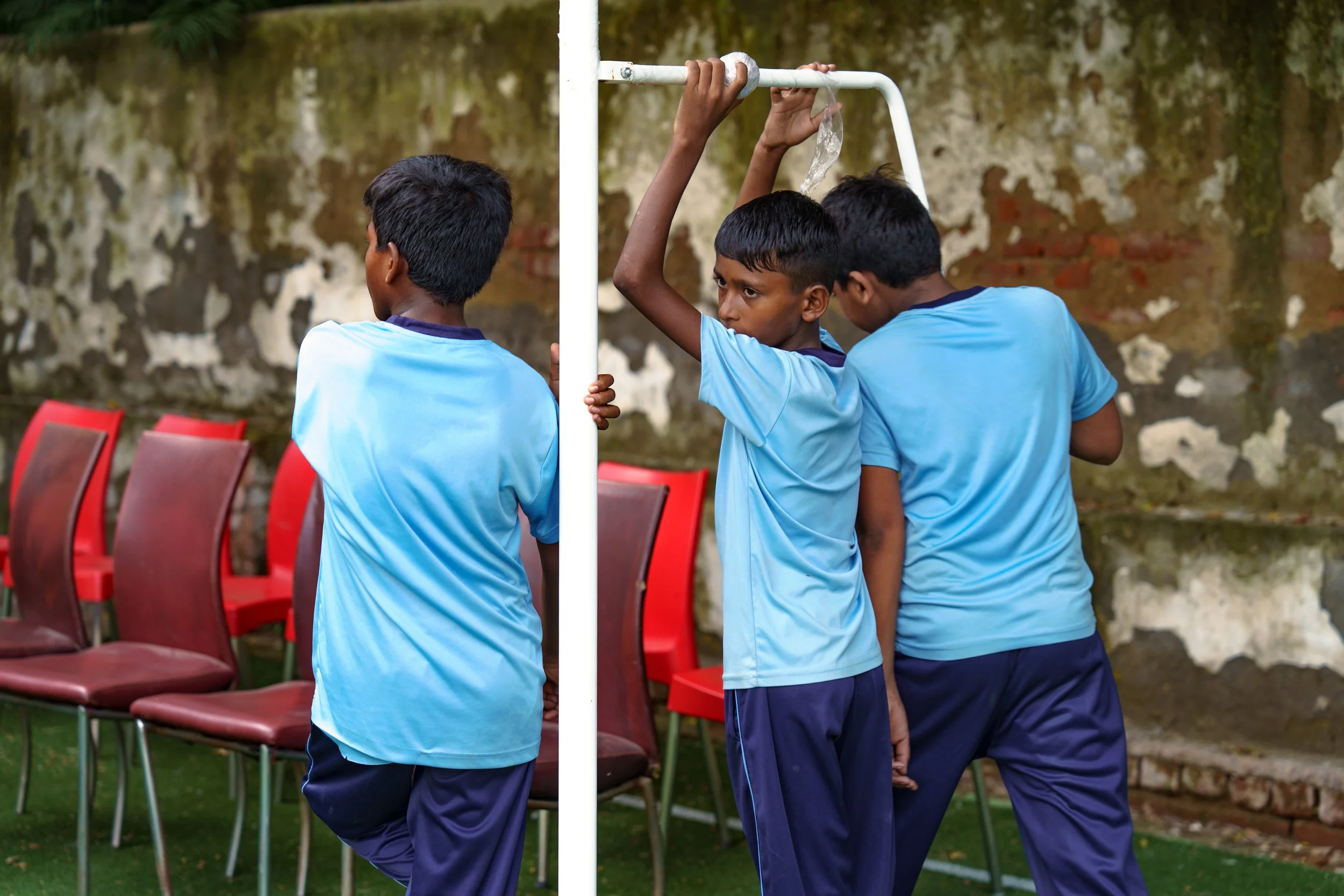 Three boys in blue shirts standing in a line on an outdoor field, participating in a game where they put a piece of cloth in their mouth to pass a white pole, with red chairs and a weathered brick wall in the background.