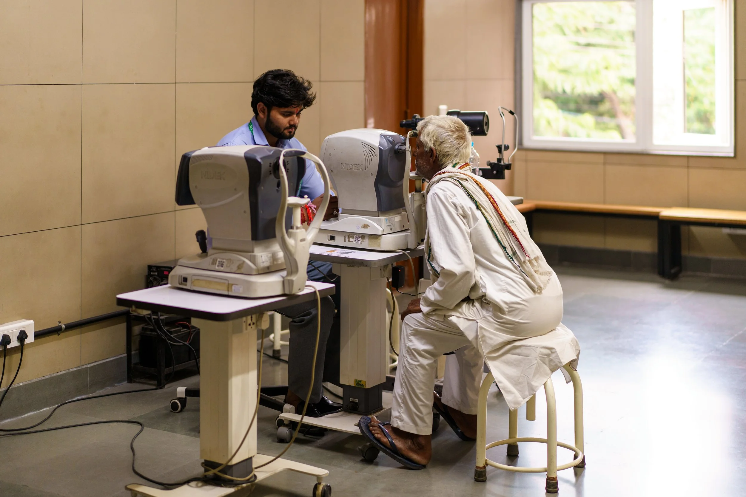 An elderly man undergoing an eye examination with an optometrist using a computer-based eye testing device in a clinic room with natural light from the open window.
