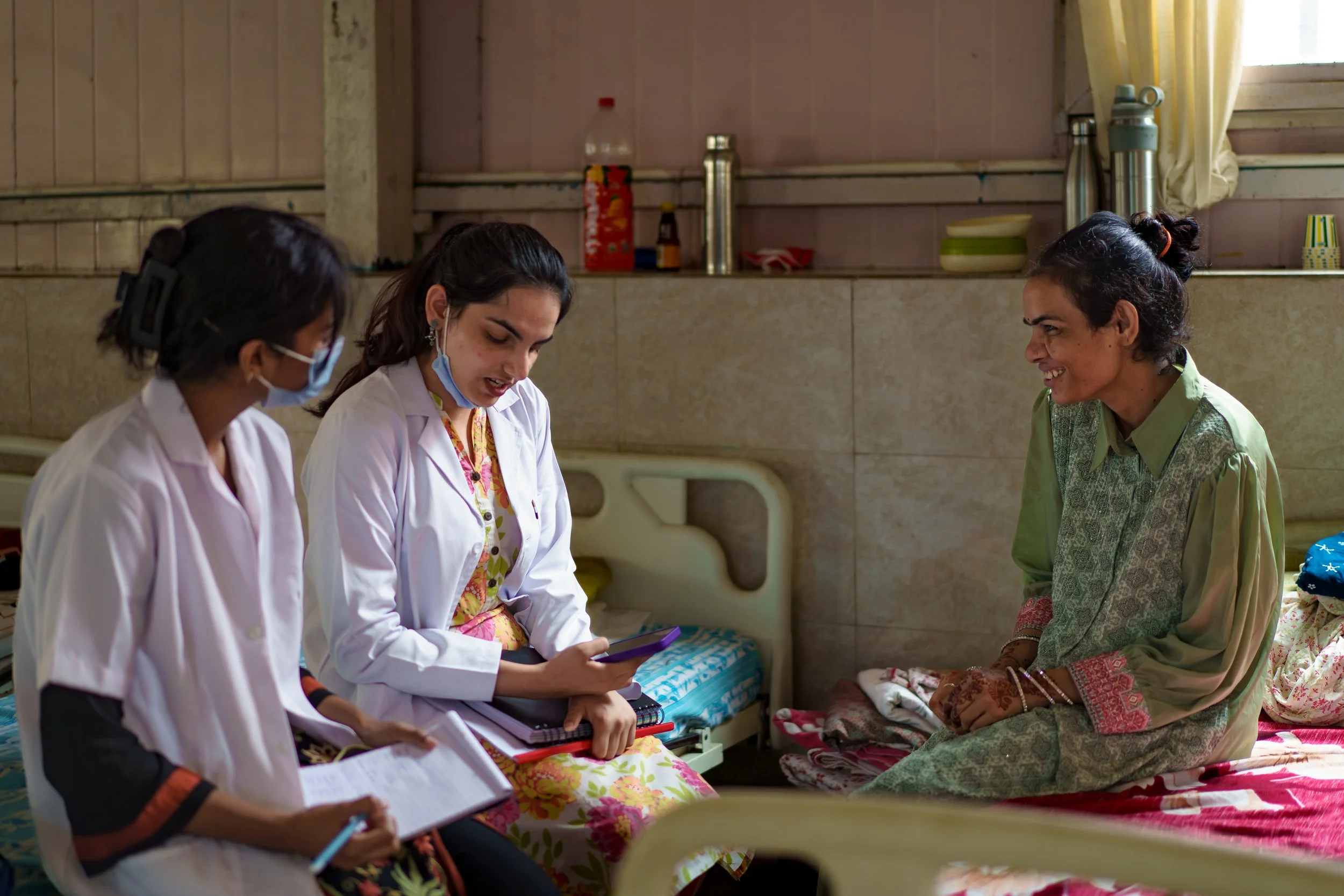 Three women, two in white medical uniforms and one in traditional clothing, are sitting on beds in a small, modest room. The women in uniforms are looking at a tablet, while the woman in traditional attire is smiling and engaging with them. The room 