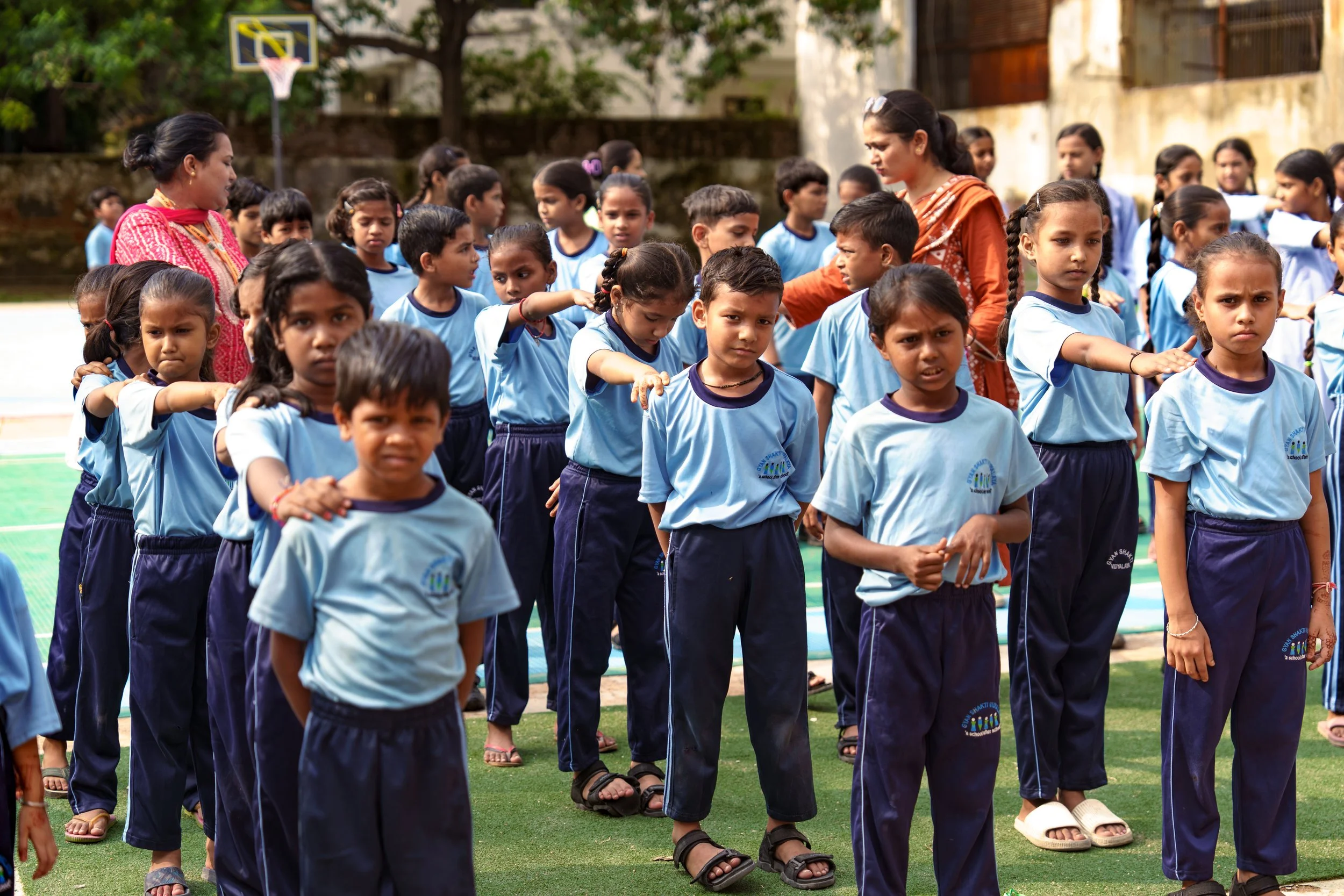 Group of school children standing outdoors on a sports field, some raising their arms, with two women supervising, a basketball hoop in the background.