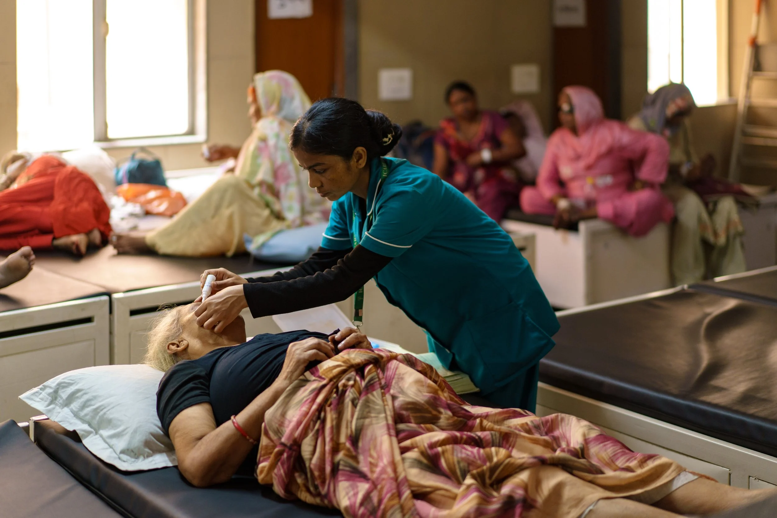 Nurse providing medical care to an elderly woman lying in a hospital bed, with other patients and medical staff in the background.
