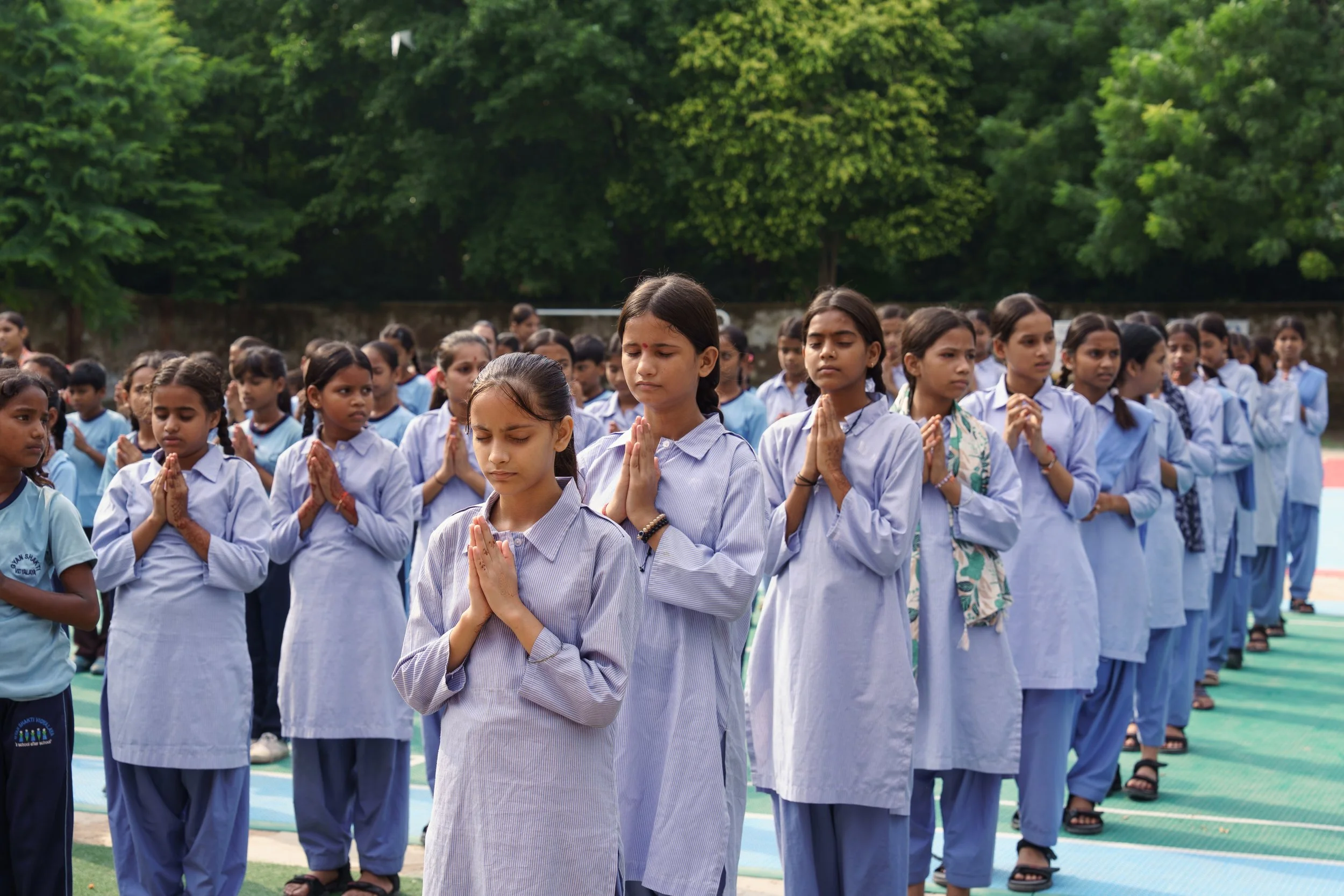 Group of young students in school uniforms practicing yoga or meditation outdoors in a line, with eyes closed and hands together in prayer position. Green trees are in the background.
