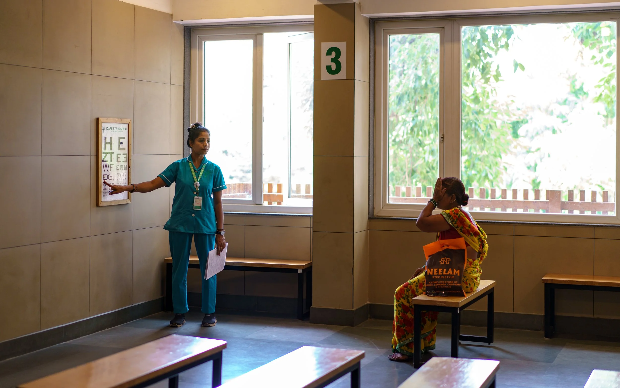A nurse in scrubs standing next to an eye chart on a wall, pointing at it, inside a clinic with two large windows, one of which frames green tree leaves outside. A woman wearing a colorful saree sits on a bench near the windows, holding her head with