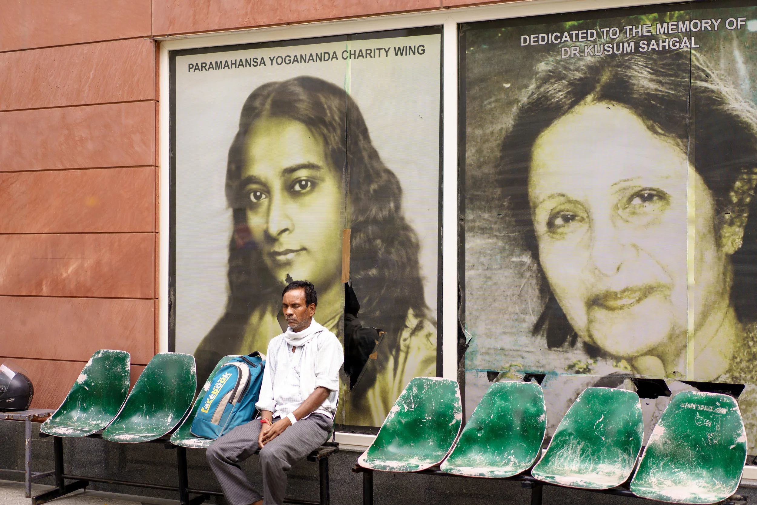 A man sitting on a green chair in front of large portrait posters of women on a building wall, with empty chairs around him.