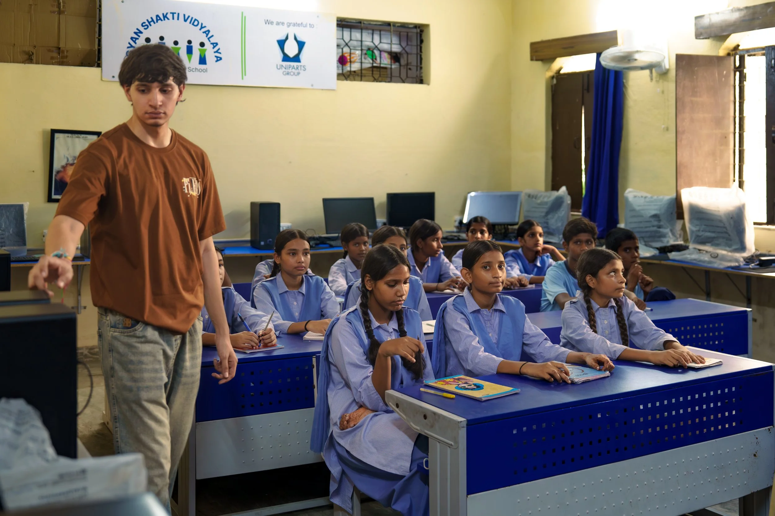 A classroom with students in blue uniforms sitting at blue desks, listening. A young man in a brown shirt is standing at the front. There are several computer monitors and a banner on the wall with school and group logos.